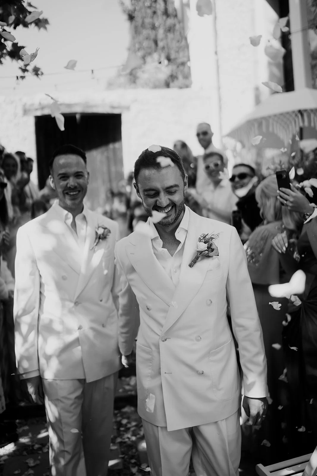 Two men in white tuxedos walking happily among a crowd at a wedding or celebration, surrounded by falling flower petals and guests taking photos.