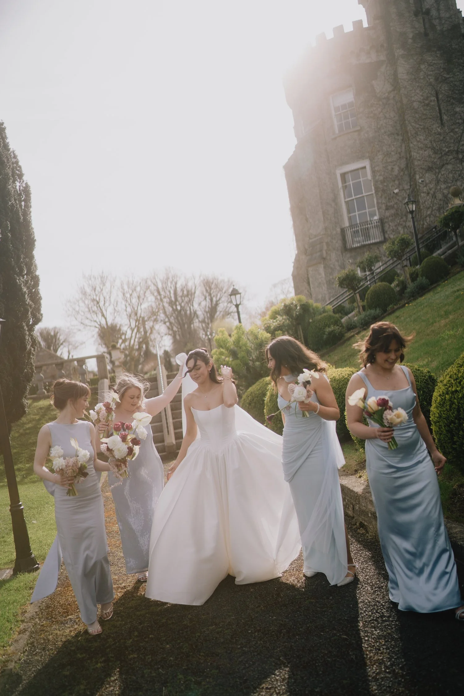 A bride and five bridesmaids walking outdoors on a sunny day, with historic stone buildings and greenery in the background. The bride is in a white wedding gown, and the bridesmaids are in light blue dresses, all carrying bouquets of flowers.