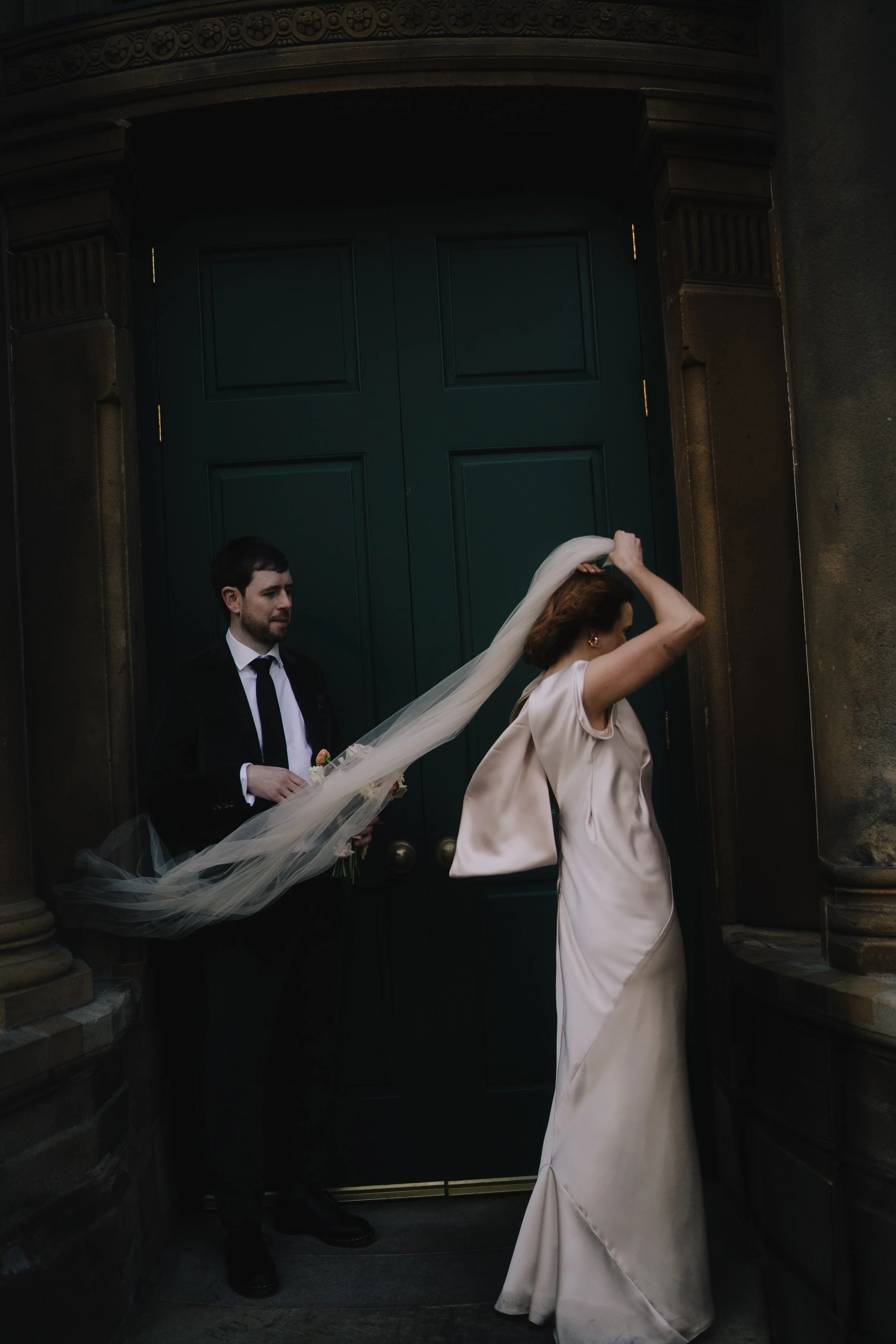 A bride in a white dress and veil adjusting her hair in front of a large dark green double door, with a groom in a black suit and tie standing nearby, holding a bouquet of flowers, outside a building with stone columns.