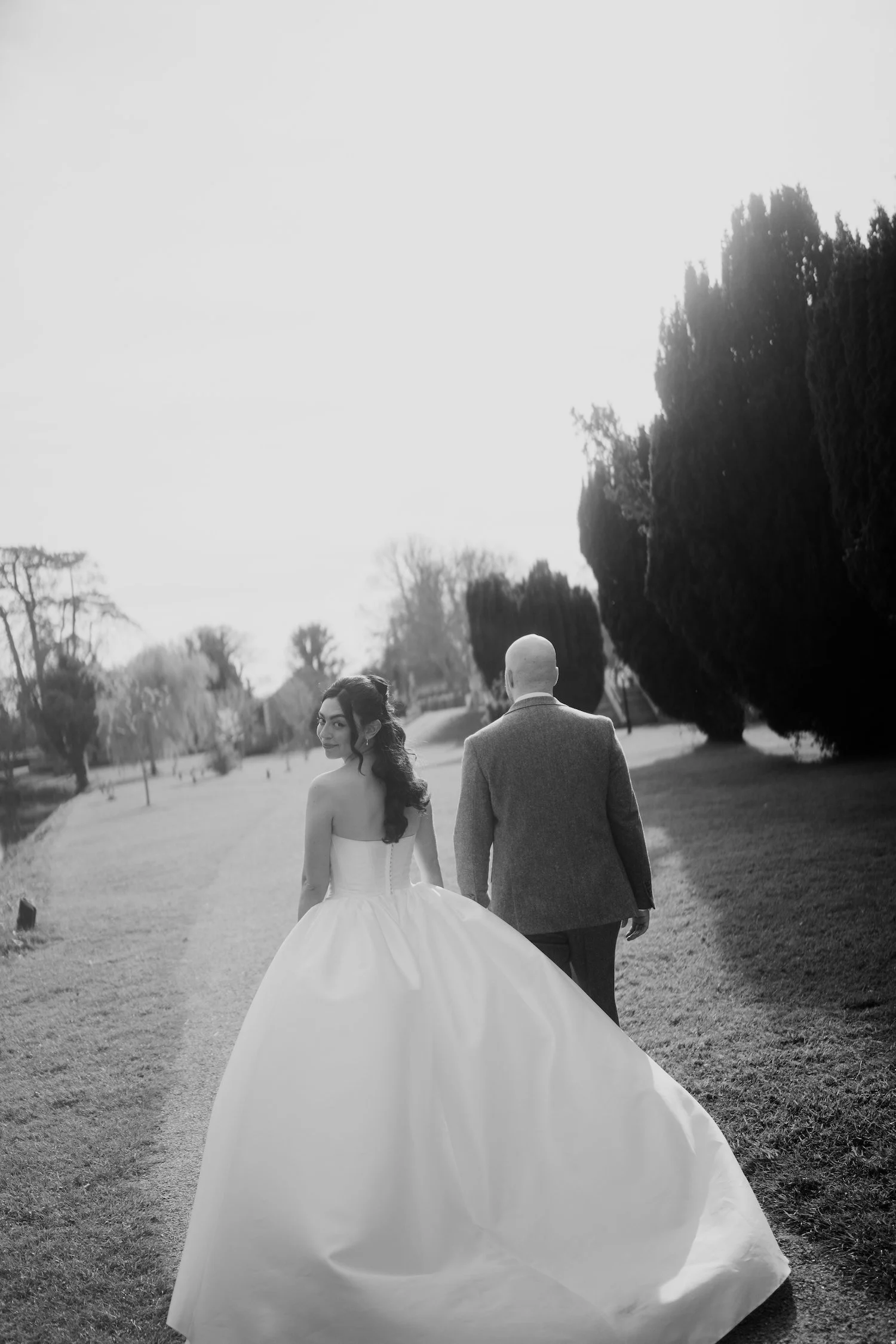 A black-and-white photo of a bride and groom walking away on a park path. The bride wears a strapless wedding gown with a long skirt, and the groom wears a suit. They are surrounded by tall trees and open grassy areas.