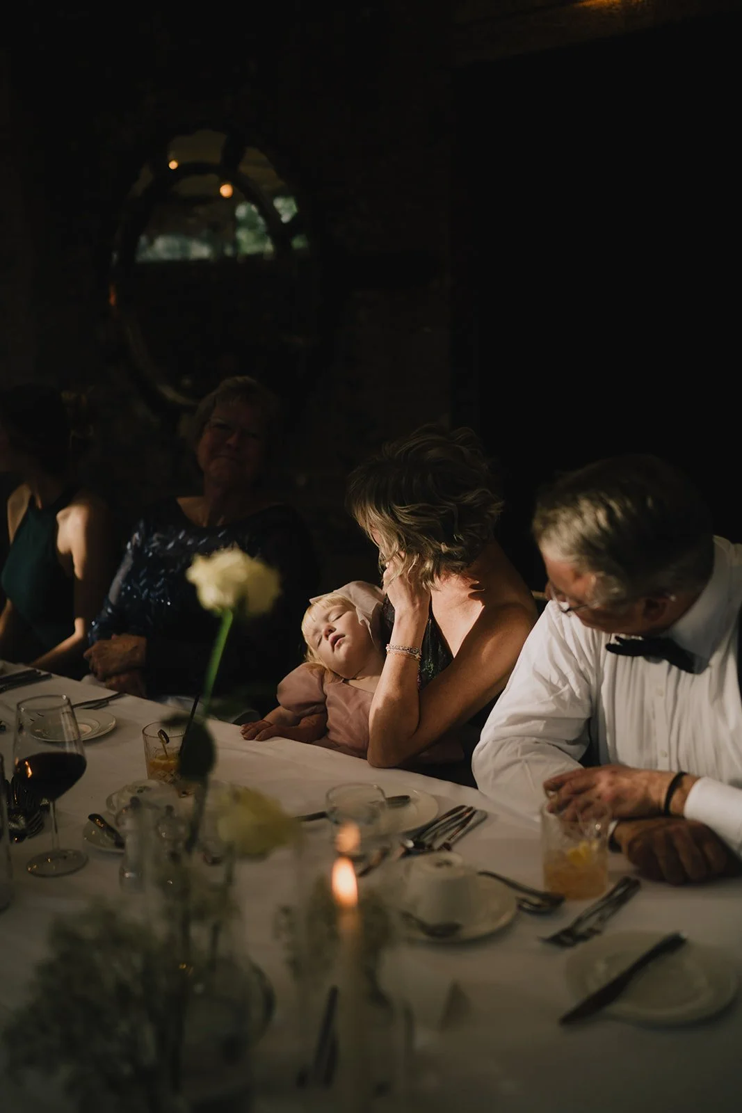 Family and friends sitting at a dinner table, with a young girl resting her head on an older woman's shoulder, in a dimly lit ambiance.