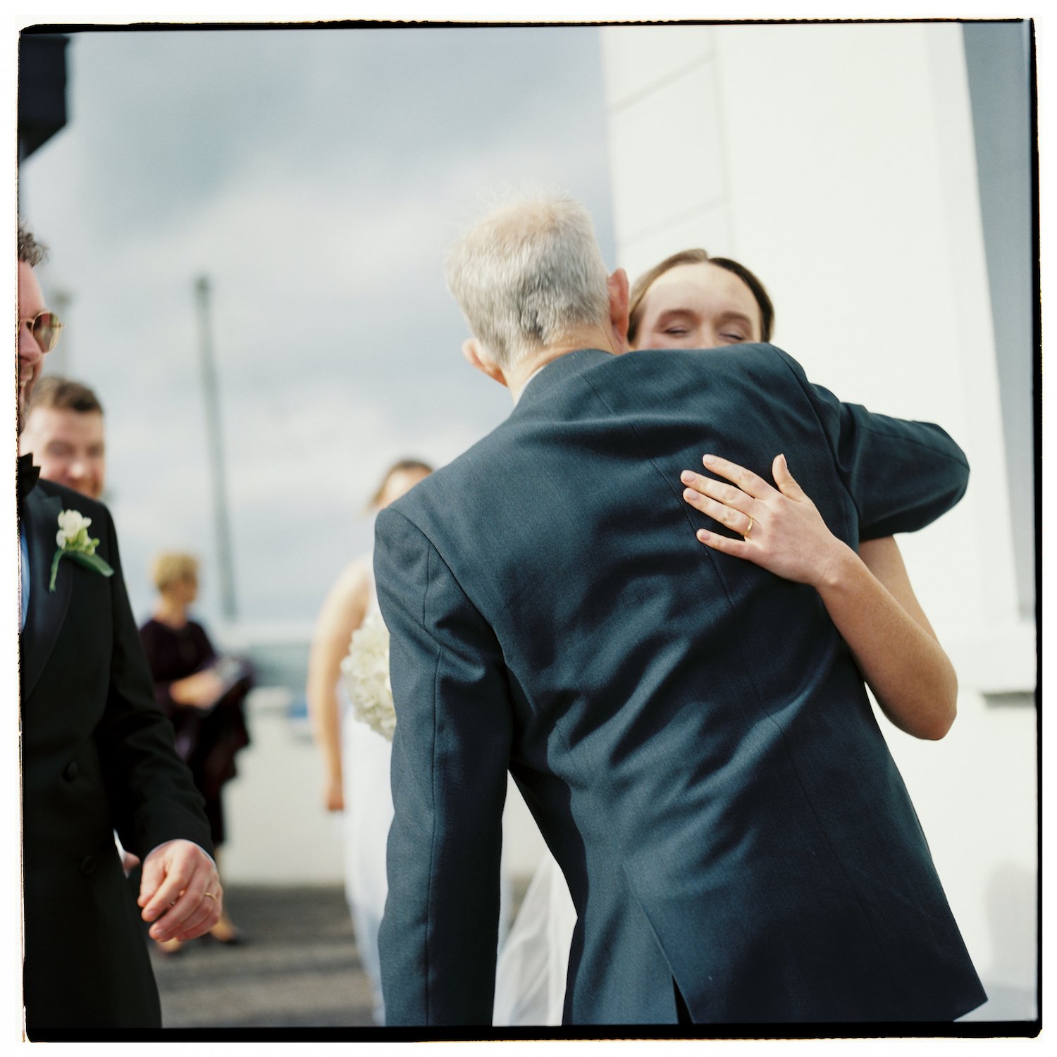 A woman in a wedding dress hugging an older man in a suit at a wedding ceremony, with a man in a tuxedo and a boutonniere in the foreground and guests in the background.