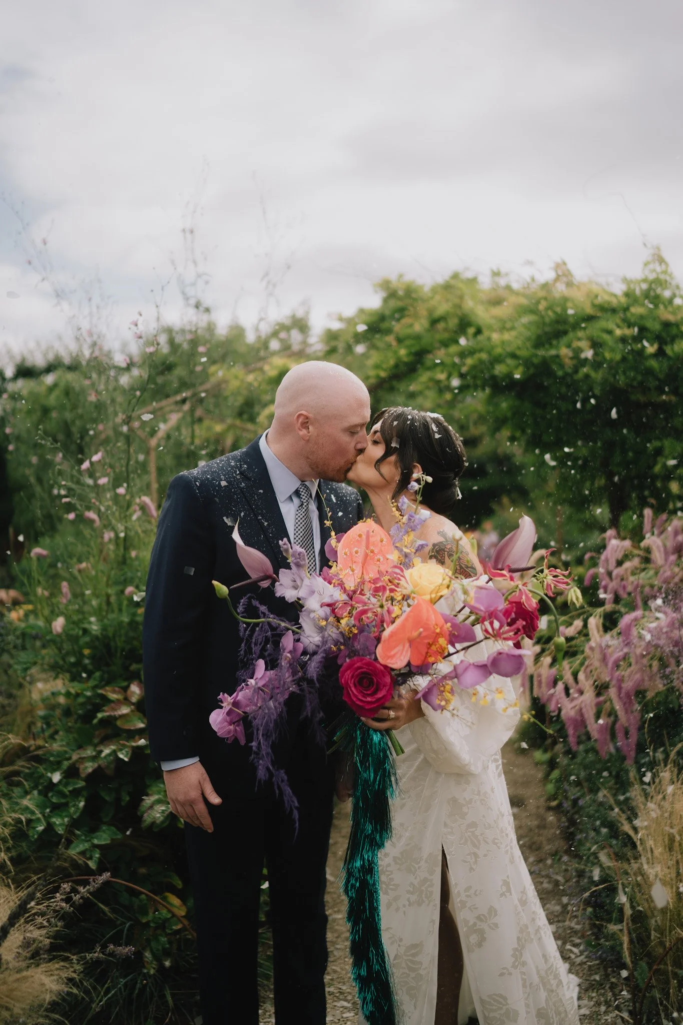 A bride and groom kiss outdoors surrounded by colorful flowers and greenery