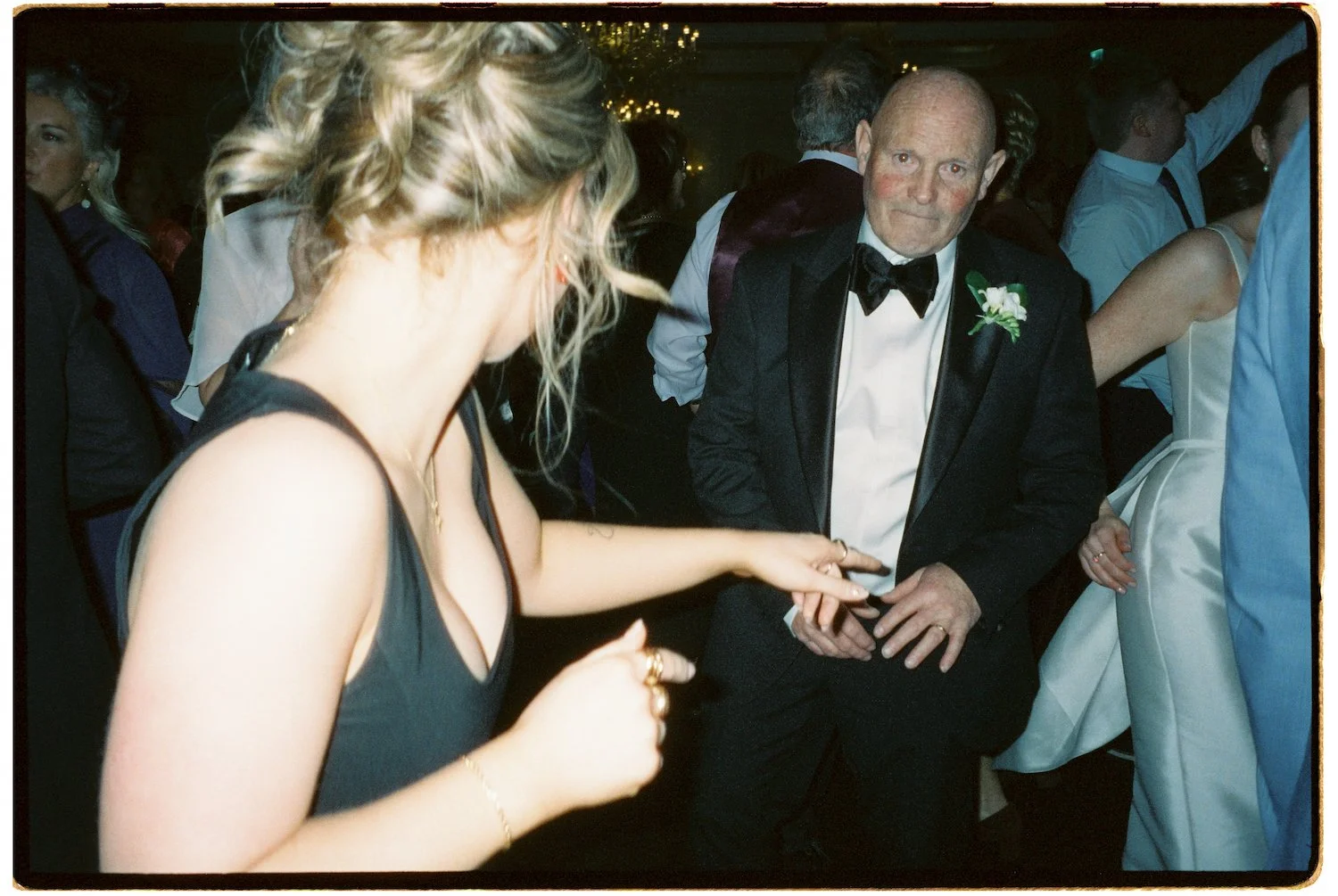 A woman in a black dress dancing with a man in a tuxedo at a formal event. Other guests are seen in the background, some dancing or socializing.