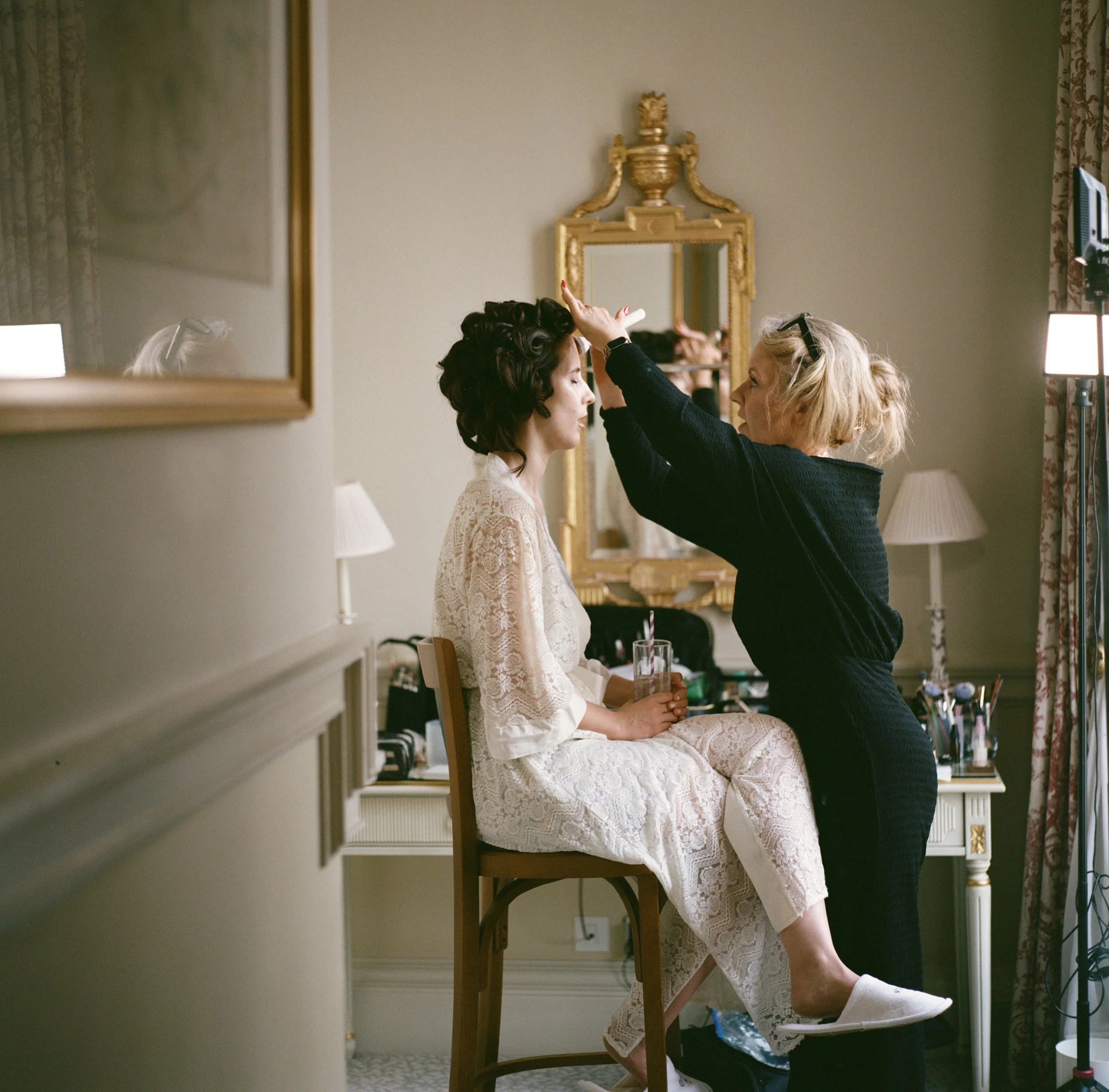 A woman with blonde hair working on a woman's hair in a room with a large ornate gold-framed mirror and floral curtains.