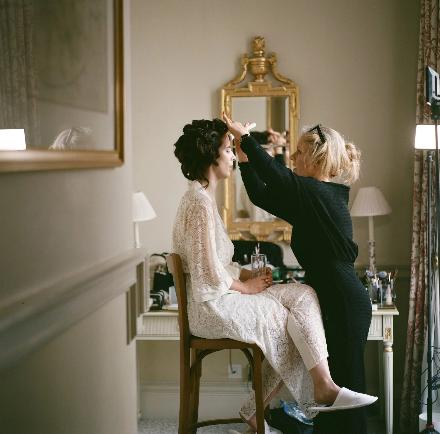 A woman in a white lace robe sits on a chair with her eyes closed while another woman in a black outfit styles her hair, in a well-lit room with gold-framed mirror and lamps.