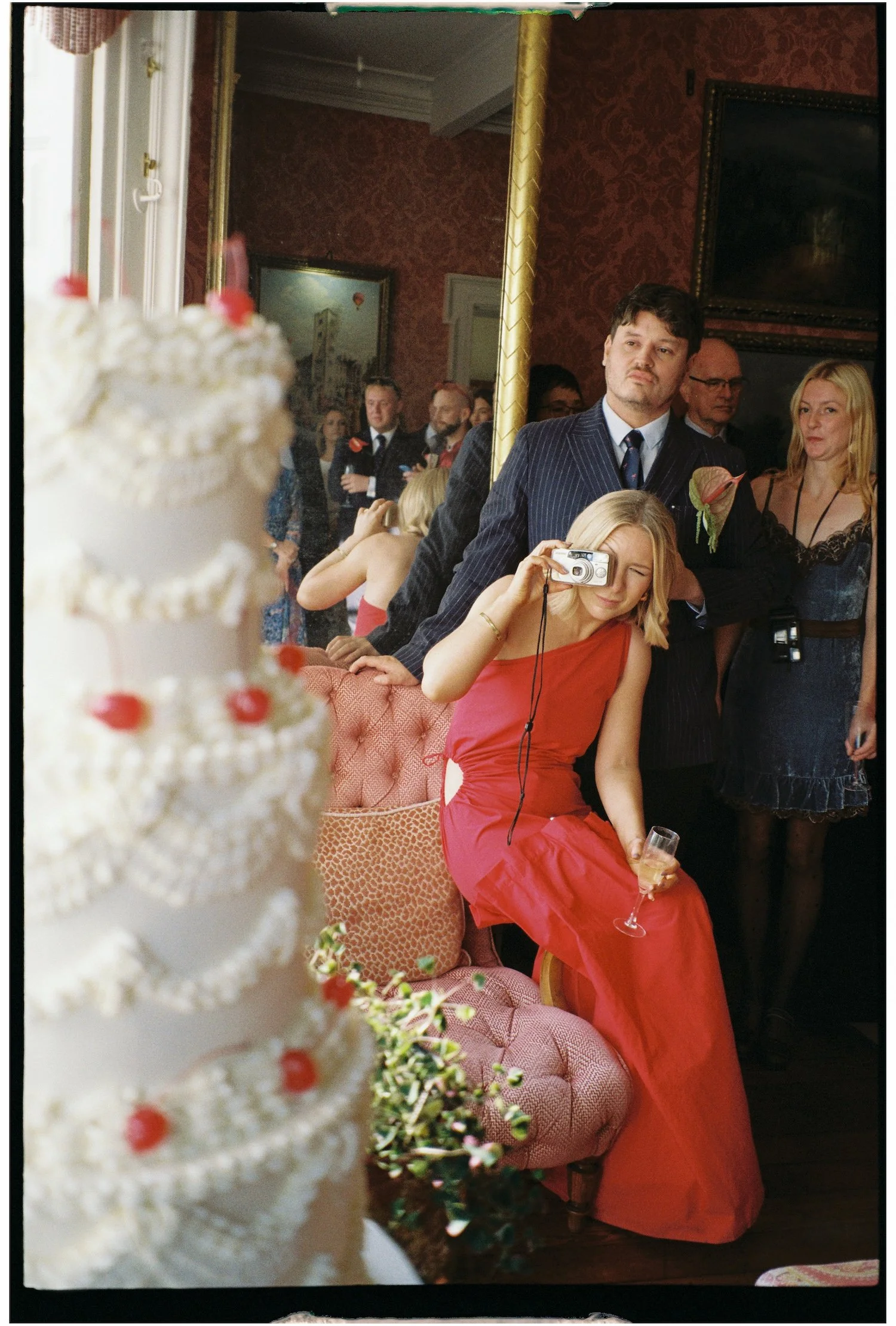 A woman in a red dress sitting on a pink velvet armchair, drinking champagne while taking a picture with a compact digital camera, at a social gathering or party with multiple people in the background.