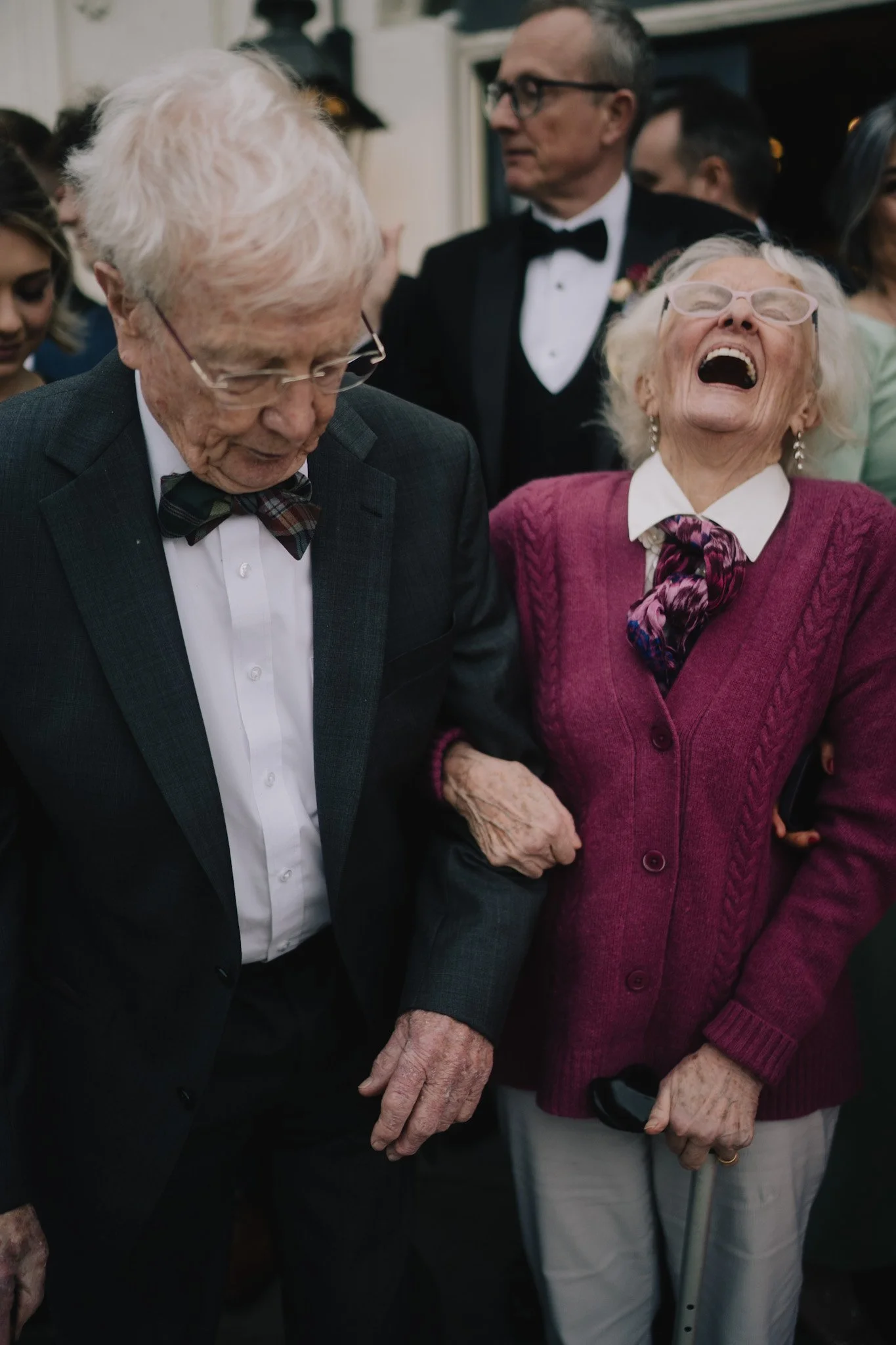 A joyful elderly woman wearing glasses and a pink sweater laughs while holding hands with an elderly man dressed in a suit and bow tie at a social gathering. The woman has white hair, and the man has white hair with glasses. There are other people in