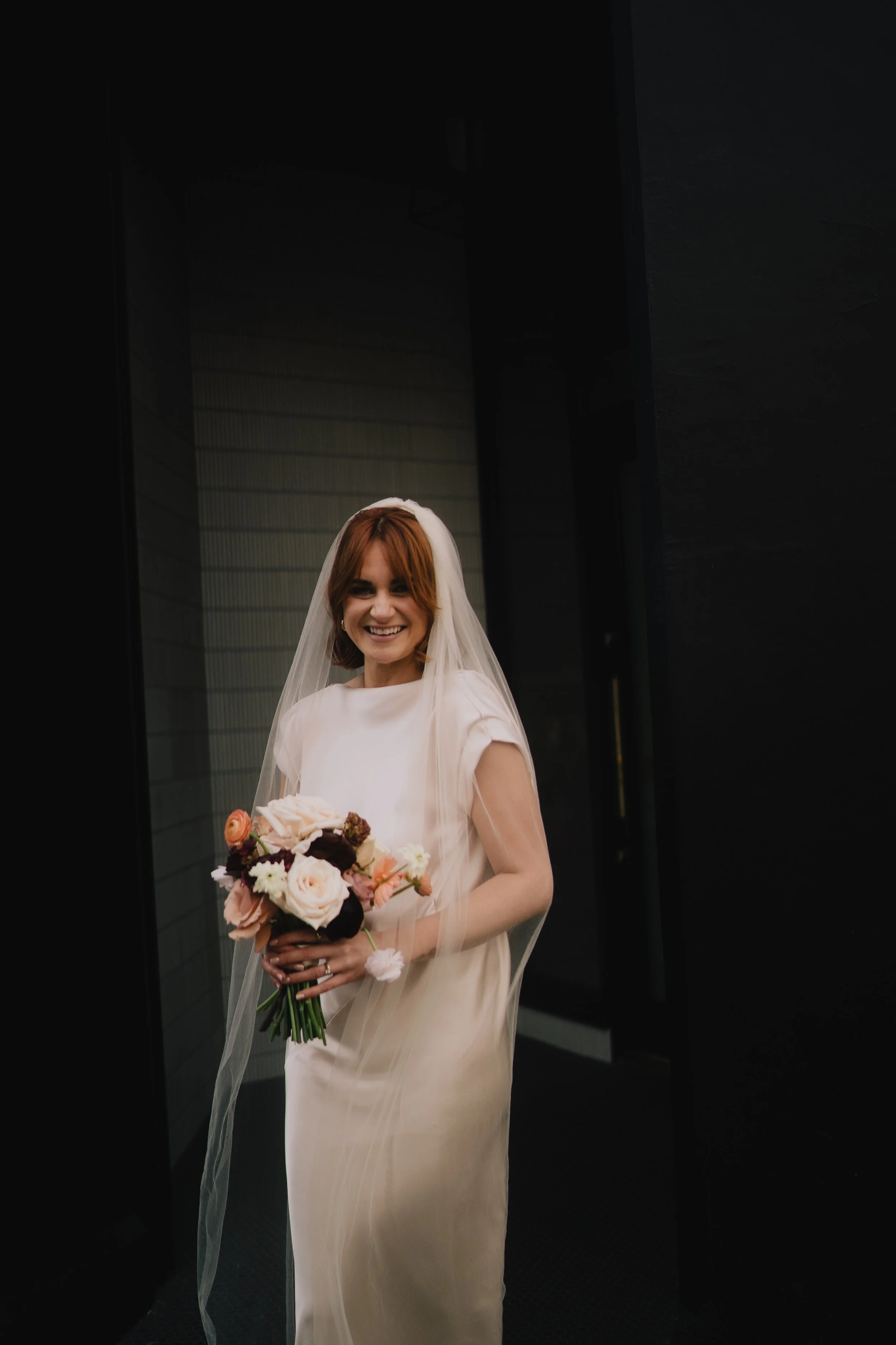 A woman dressed as a bride in a light-colored wedding gown with short sleeves, holding a bouquet of pale pink, cream, and dark red flowers, with a long veil on her red hair, smiling in front of a dark background.
