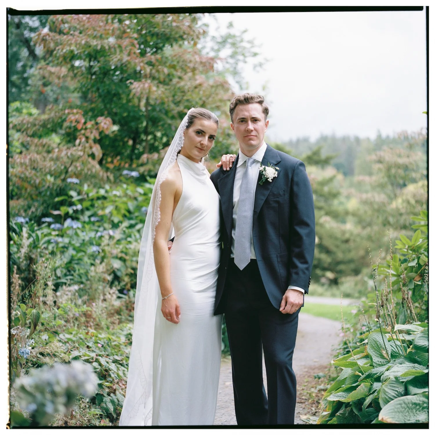 A bride and groom standing outdoors, surrounded by greenery and flowering plants, posing for a wedding photo. The bride is wearing a white wedding dress and veil, and the groom in a dark suit with a boutonniere.
