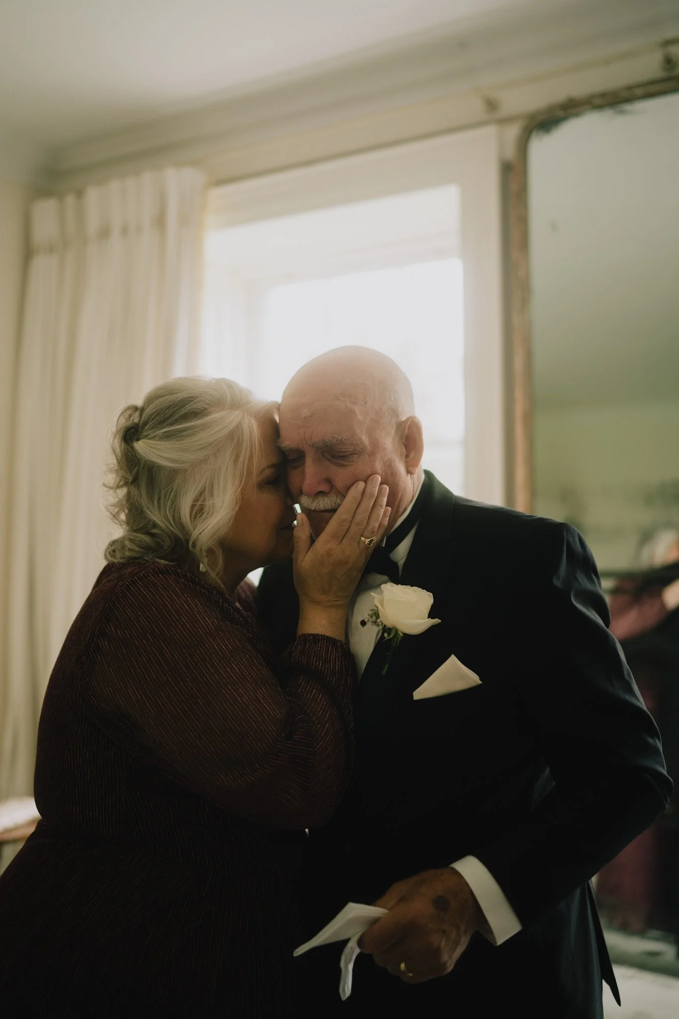 An elderly woman and an elderly man share an intimate moment at a formal event, possibly a wedding. The woman gently touches the man's face while they lean their heads close together, showing affection amidst a softly lit setting.
