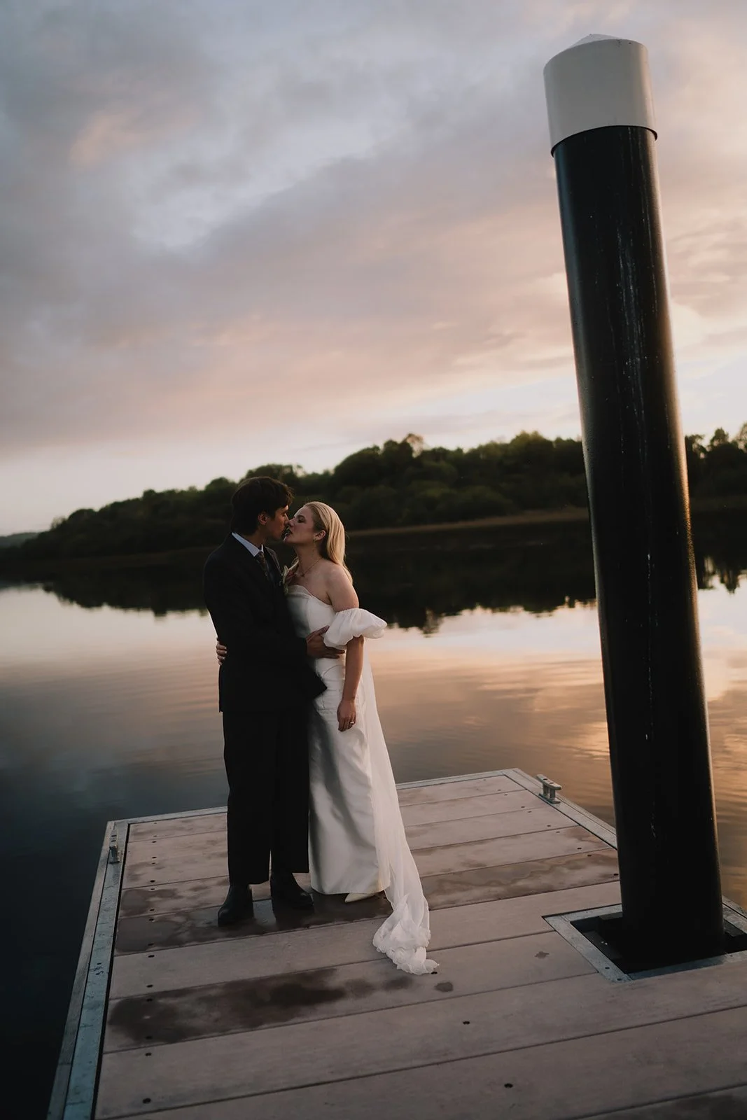 A couple dressed in wedding attire sharing a kiss on a dock by a lake at sunset.