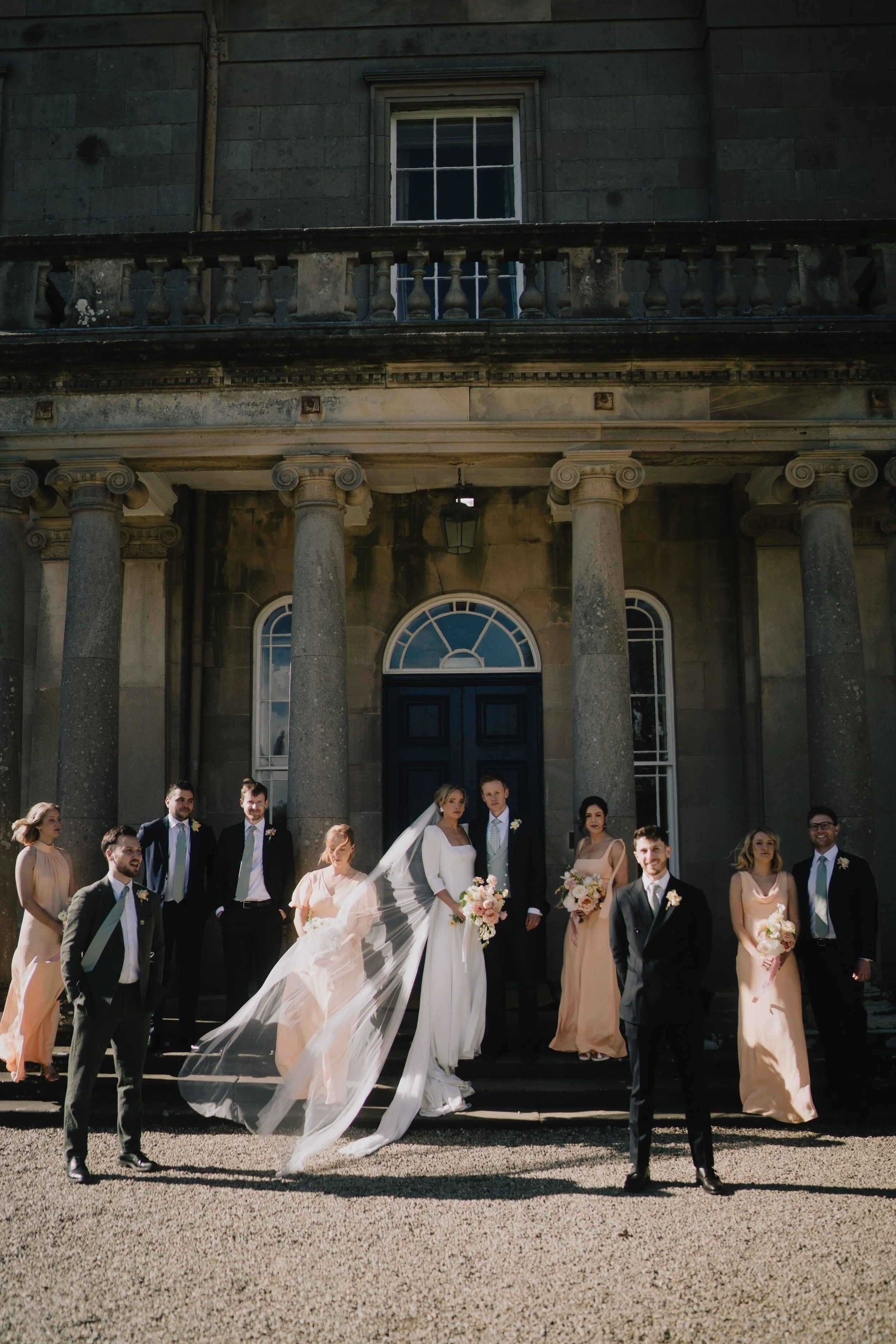 Wedding party standing in front of a historic stone building with columns and large windows. The bride and groom are centered, with the bride in a white dress and veil holding a bouquet, and the groom in a dark suit with a boutonniere. Bridesmaids in