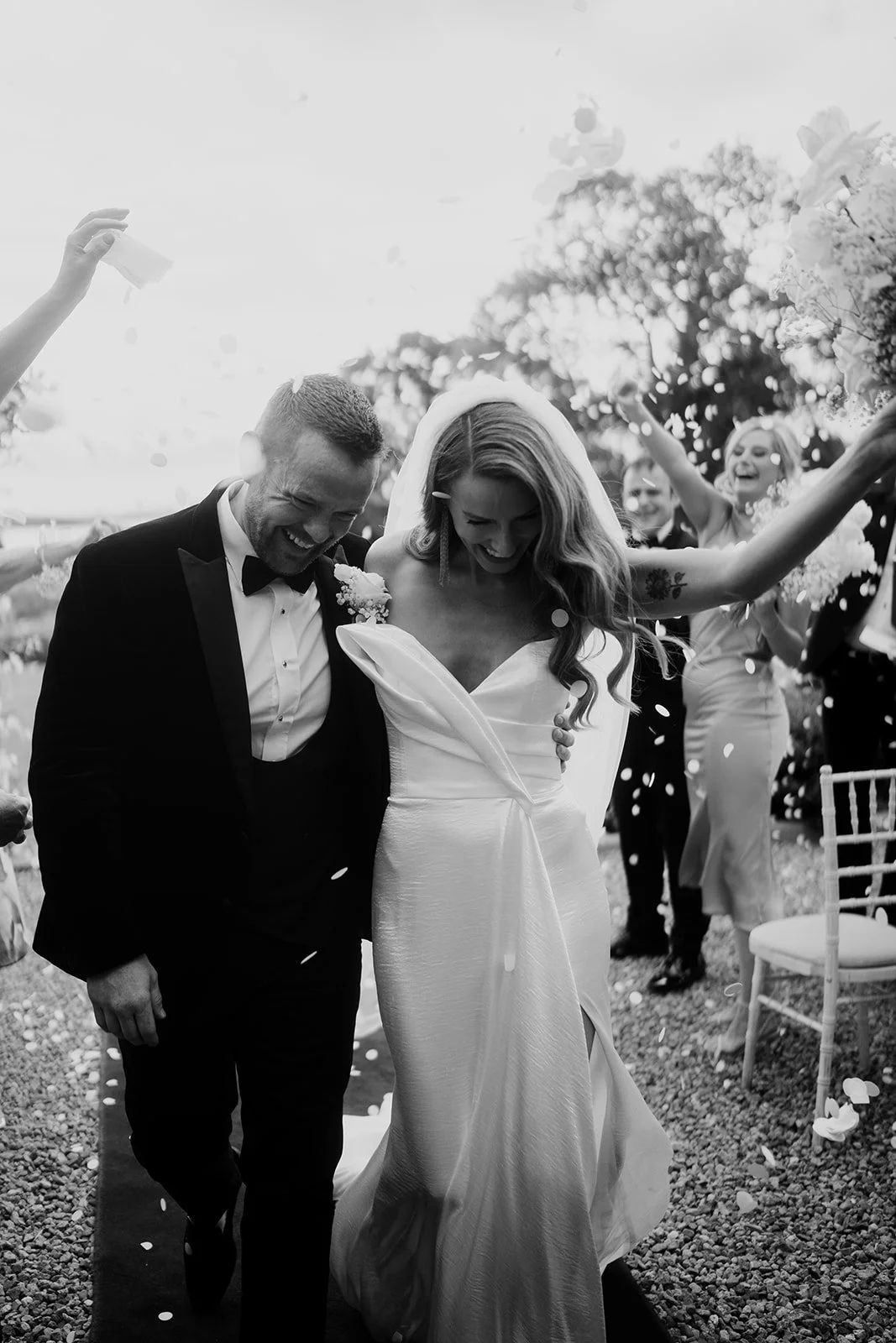 Black and white photo of a wedding celebration with a bride and groom smiling and walking together, surrounded by friends throwing confetti.