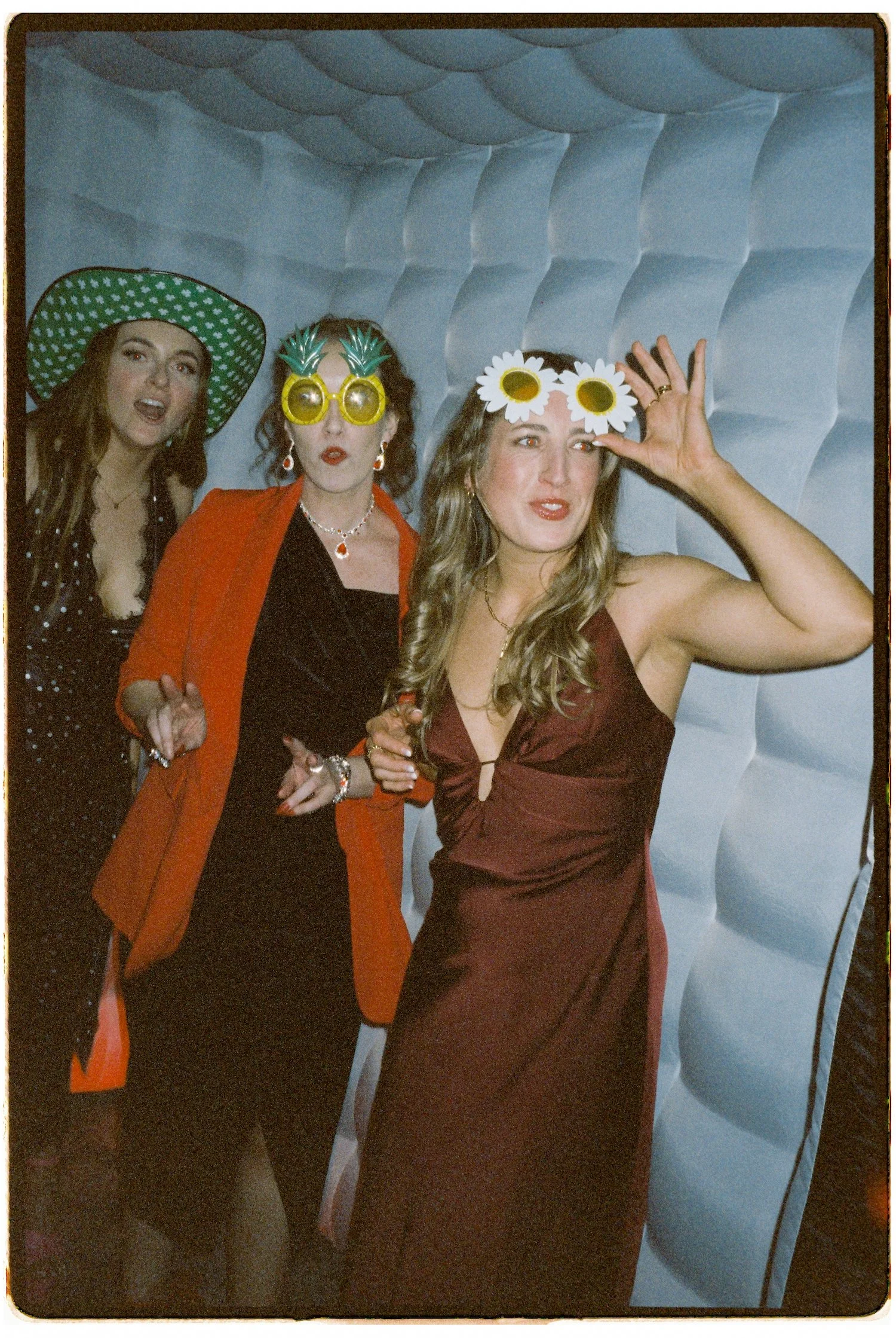 Three women posing in a photo booth, wearing colorful sunglasses and fun accessories, with a blue padded wall background.