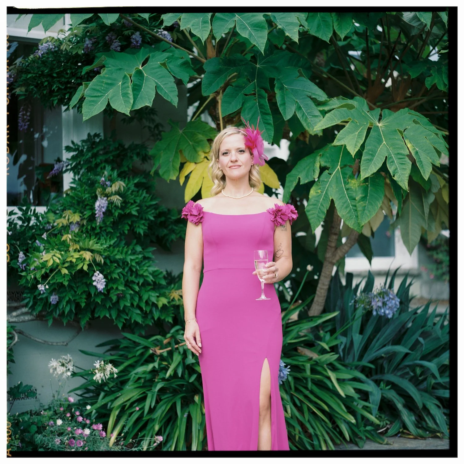A woman in a pink evening gown with floral shoulder accents and a slit in the dress, holding a glass of champagne, standing in front of lush green plants and large leafy trees.