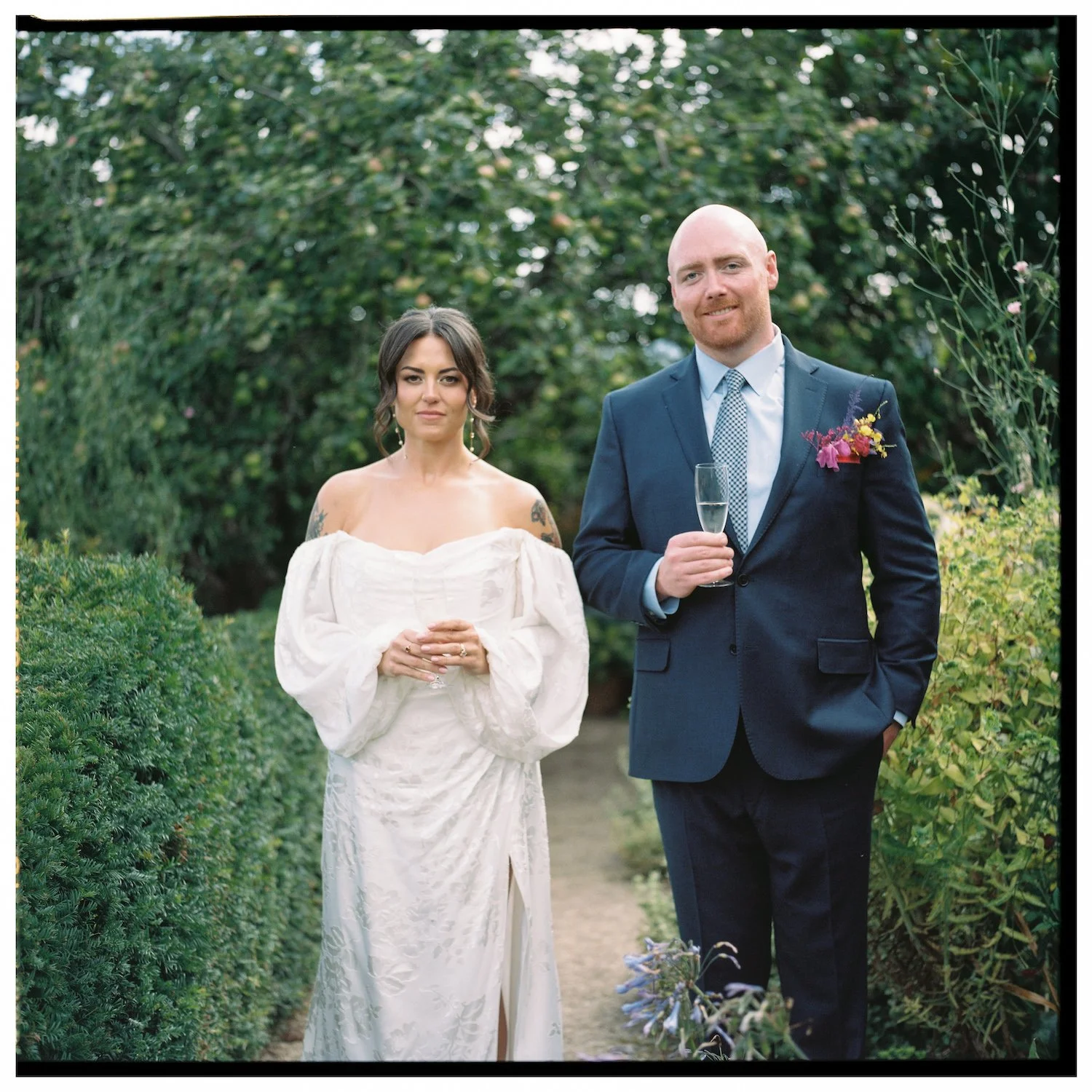 A bride and groom standing outdoors in a garden, both holding glasses and dressed in wedding attire, with trees and bushes surrounding them.