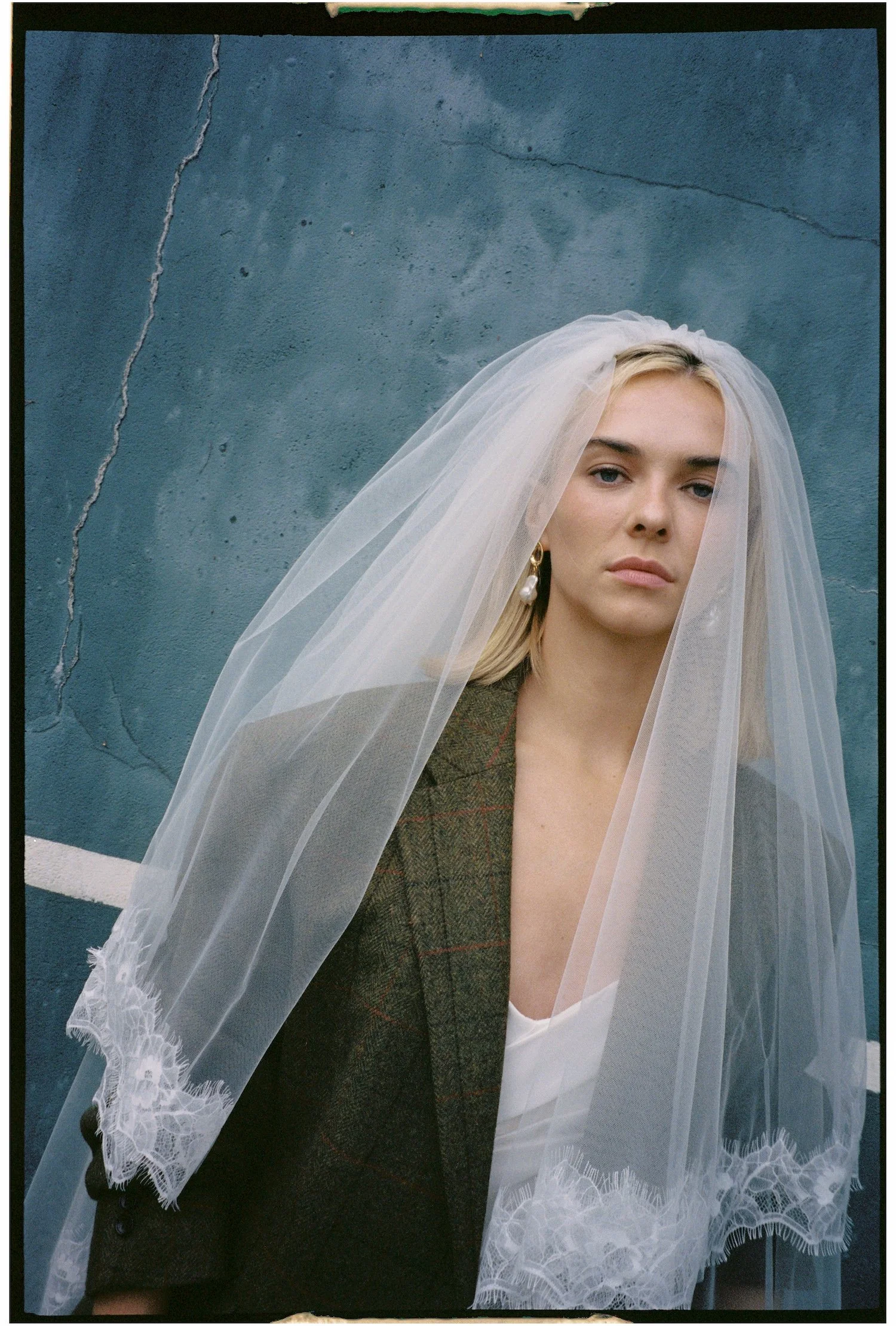 Close-up of a woman wearing a wedding veil with lace edge, wearing earrings, against a blue wall with cracks. She has light blonde hair, and a serious expression.