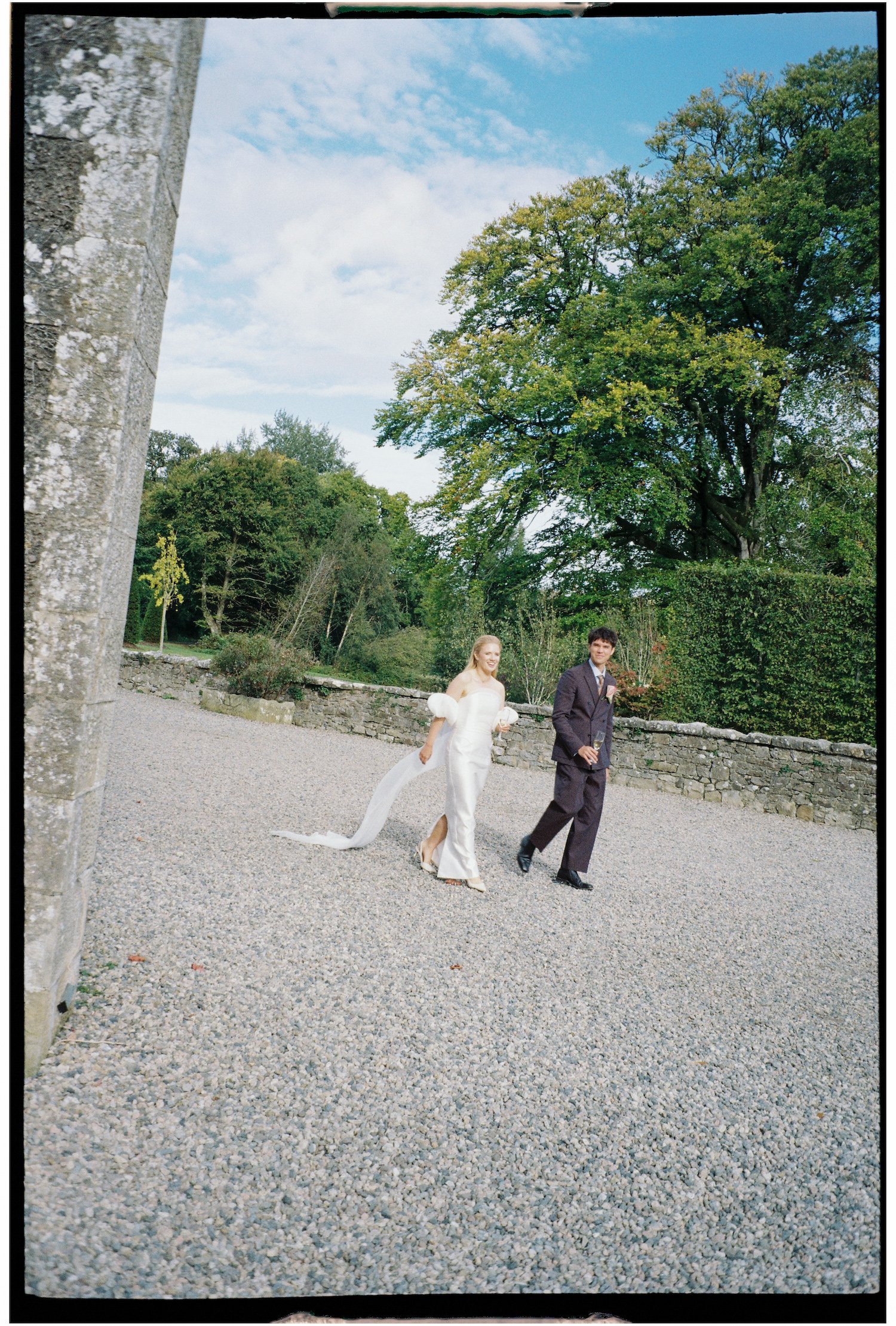 A bride and groom walking outside on a gravel path in a park-like setting during daytime, with trees and a stone wall in the background.