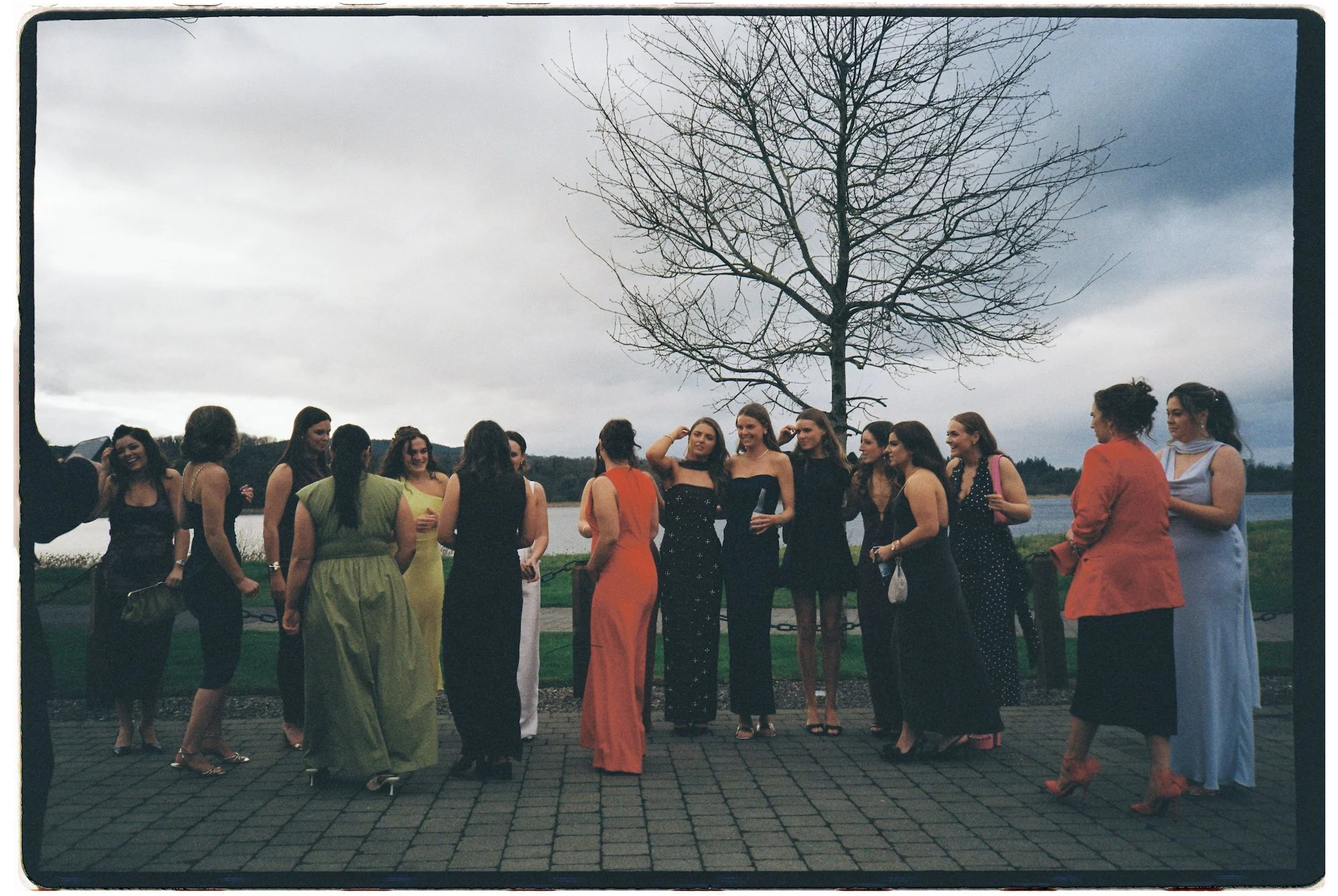 A group of women standing together outdoors on a paved area near a body of water, with a large, leafless tree and cloudy sky in the background. They are dressed in colorful, elegant dresses, possibly for a formal event or celebration.