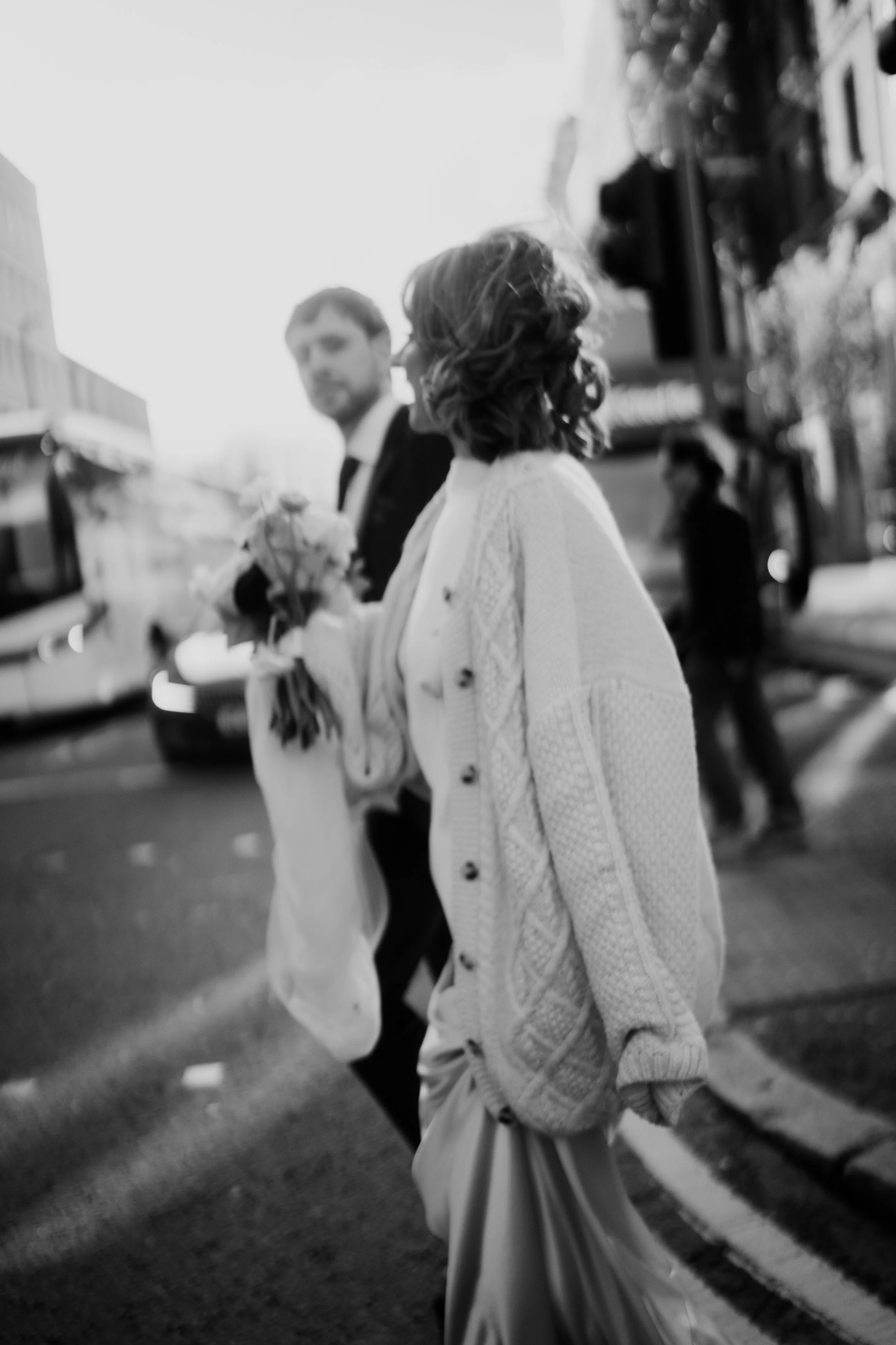 Black and white photo of two women walking across a city street, one holding a bouquet of flowers, with buildings and a traffic light in the background.