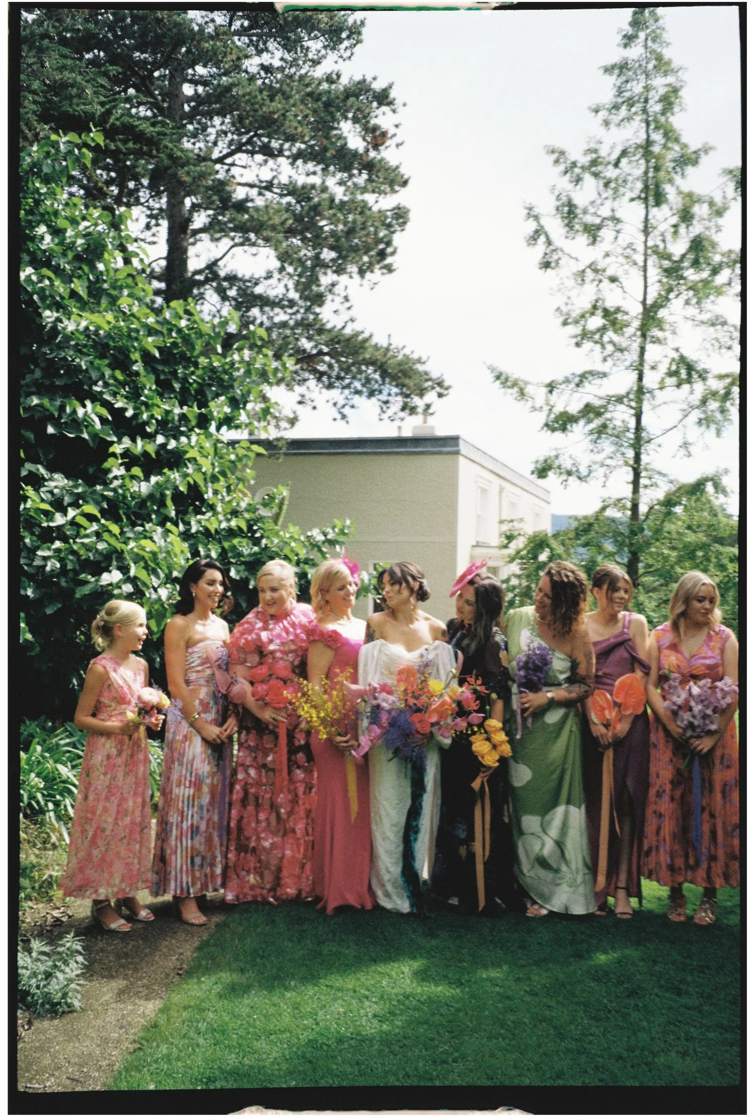 A group of women wearing colorful dresses, some holding flowers, standing outdoors in a garden.
