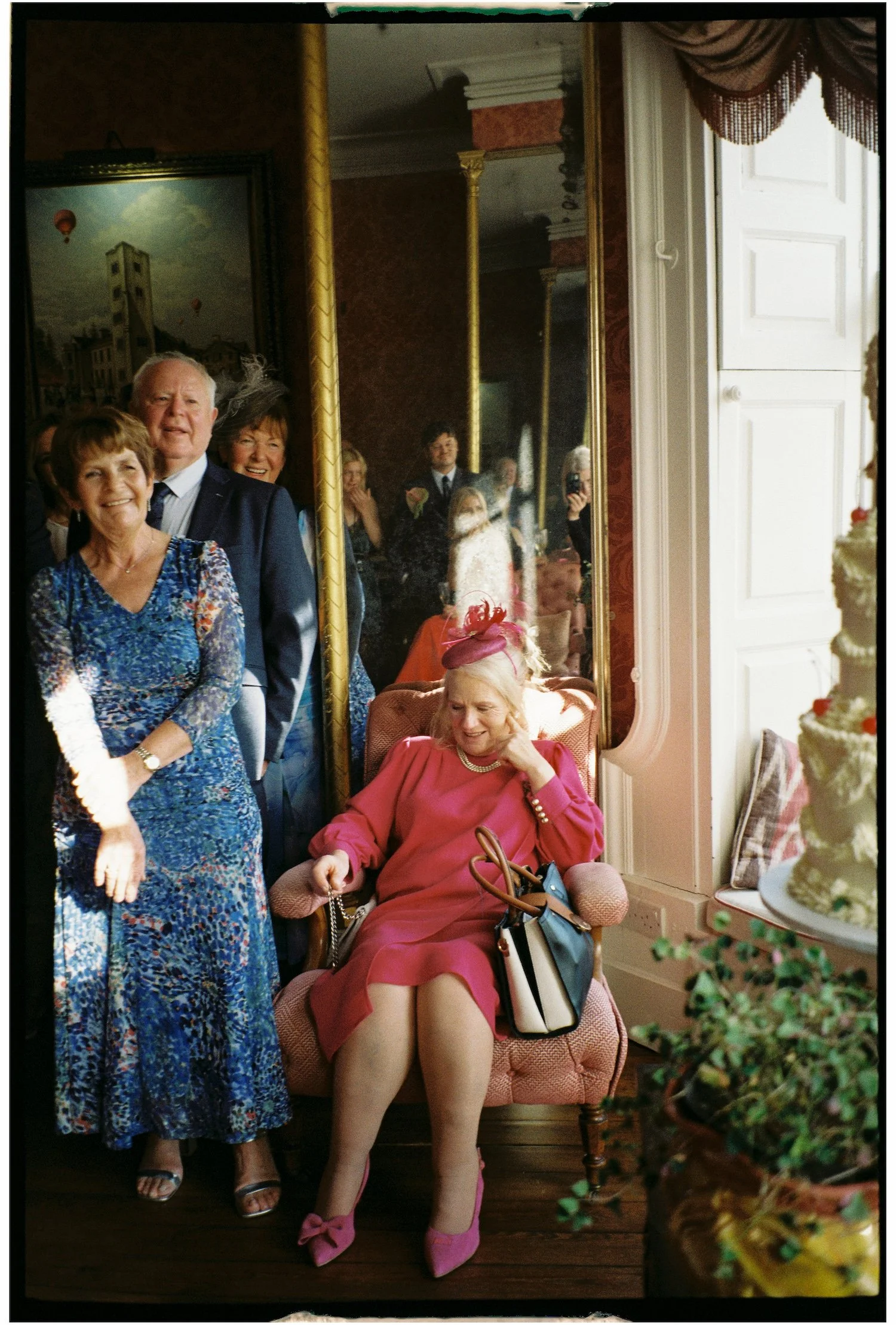 A group of elderly adults gathered in a warmly decorated room, with one woman seated on a pink velvet armchair, dressed in pink with matching shoes, surrounded by standing companions smiling and posing for a photo.