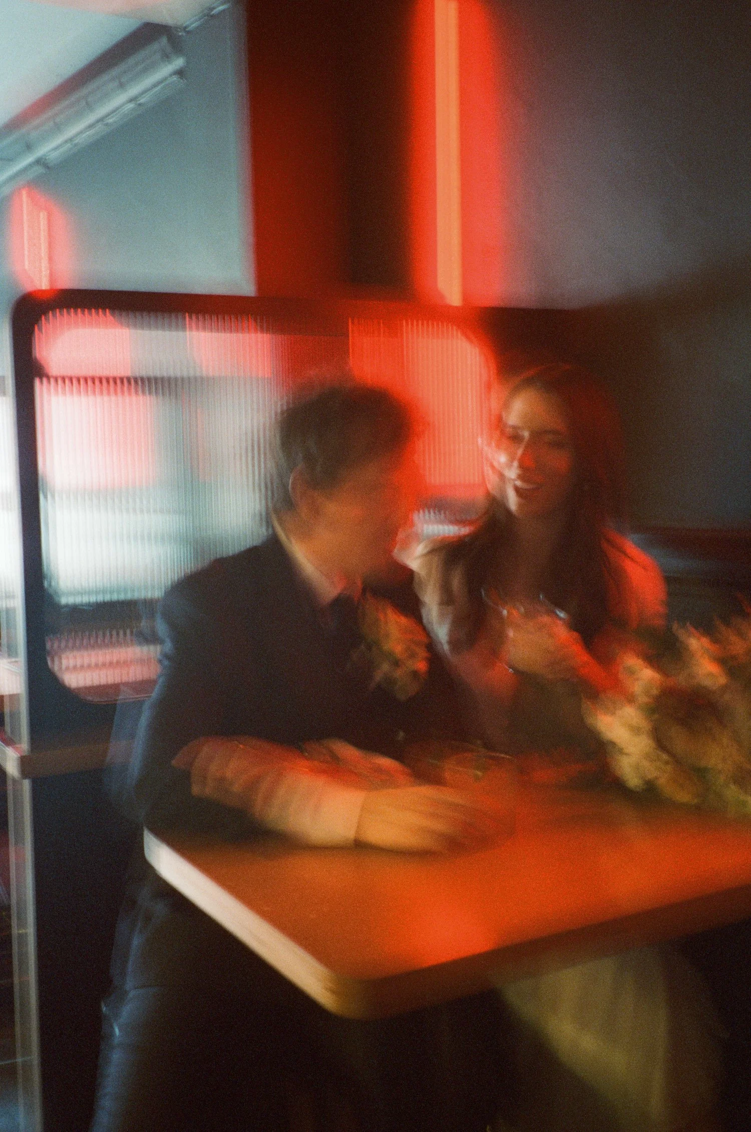 Two people, a man in a dark suit and a woman in a light-colored dress, are sitting at a table with a large flower arrangement. They are smiling and appear to be engaged in conversation. The background has a red and gray illuminated design.