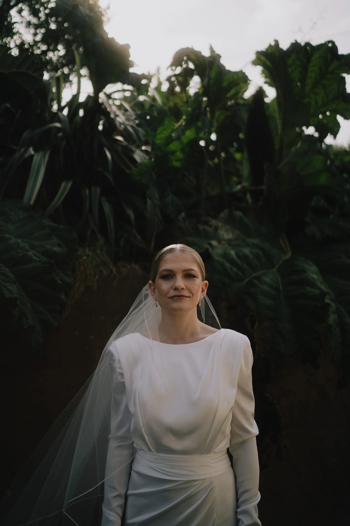 A woman in a white wedding dress and veil standing outdoors surrounded by large green plants, with sunlight behind her.