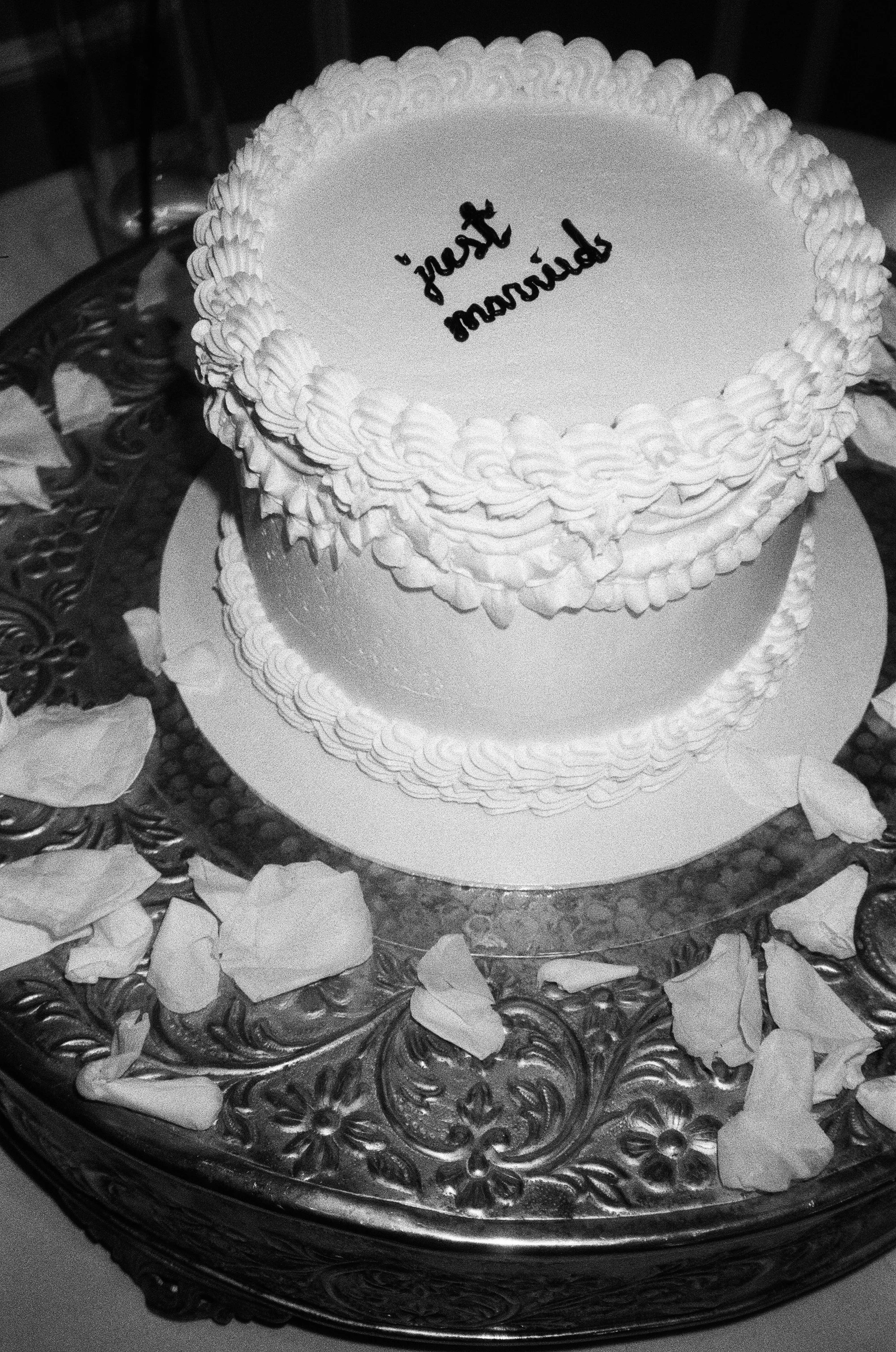 A round cake with white frosting and piped borders on top. The cake has text written in black icing that appears to say 'Happy Birthday' but is upside down. The cake is placed on an ornate metal tray surrounded by flower petals.