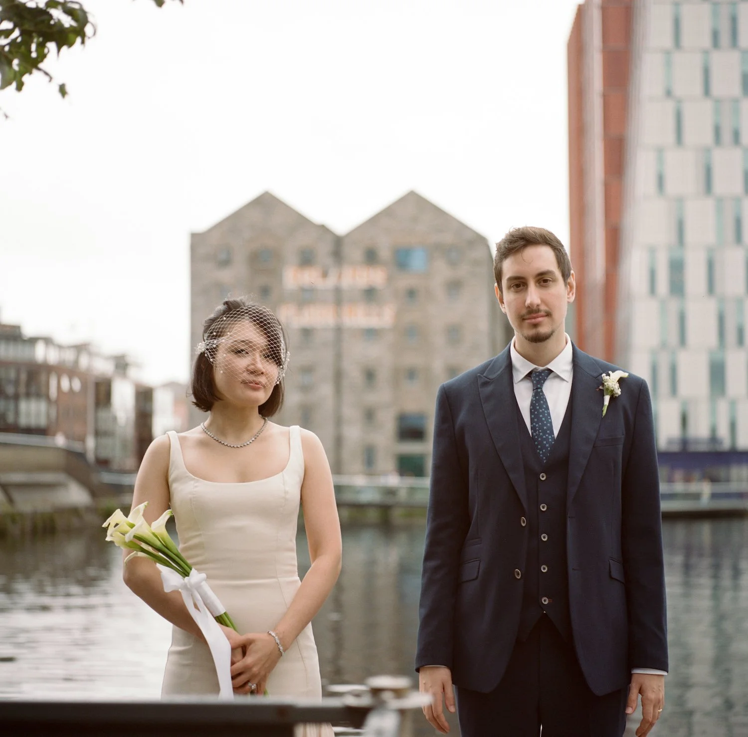 A bride and groom standing outdoors by a river with urban buildings in the background. The bride is holding a bouquet of white calla lilies and wearing a white dress with a birdcage veil. The groom is wearing a navy suit with a boutonnière. Both are 