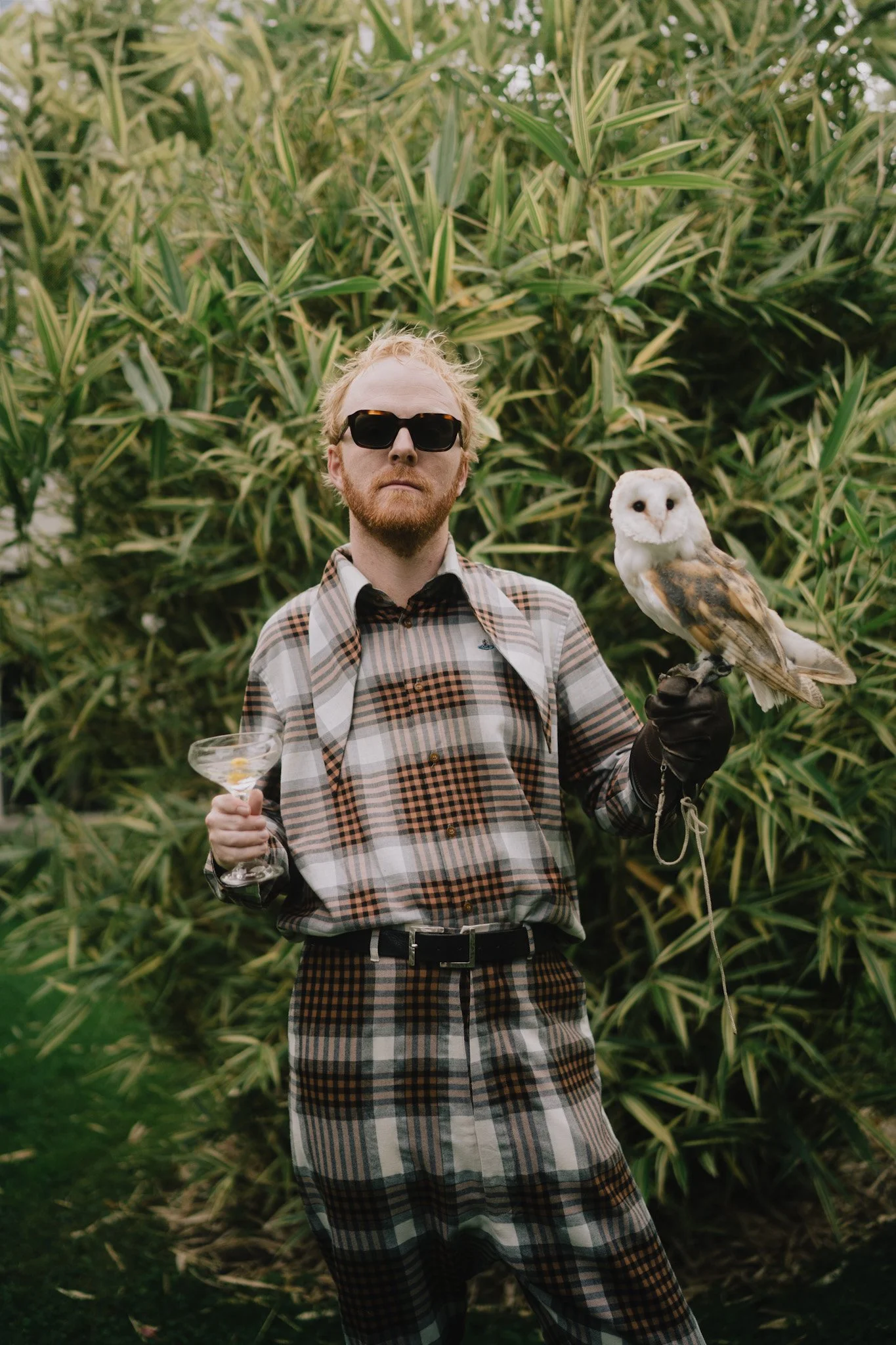 A man with blonde hair, a beard, wearing black sunglasses, a plaid shirt, and matching plaid pants, holding a glass of drink in one hand and an owl on his gloved hand, standing outdoors in front of overgrown greenery.