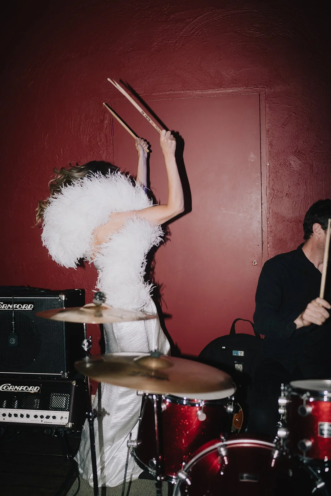 A woman in a white feathered dress is playing drums on a stage with a red wall in the background, and a man in black is playing musical instruments beside her.