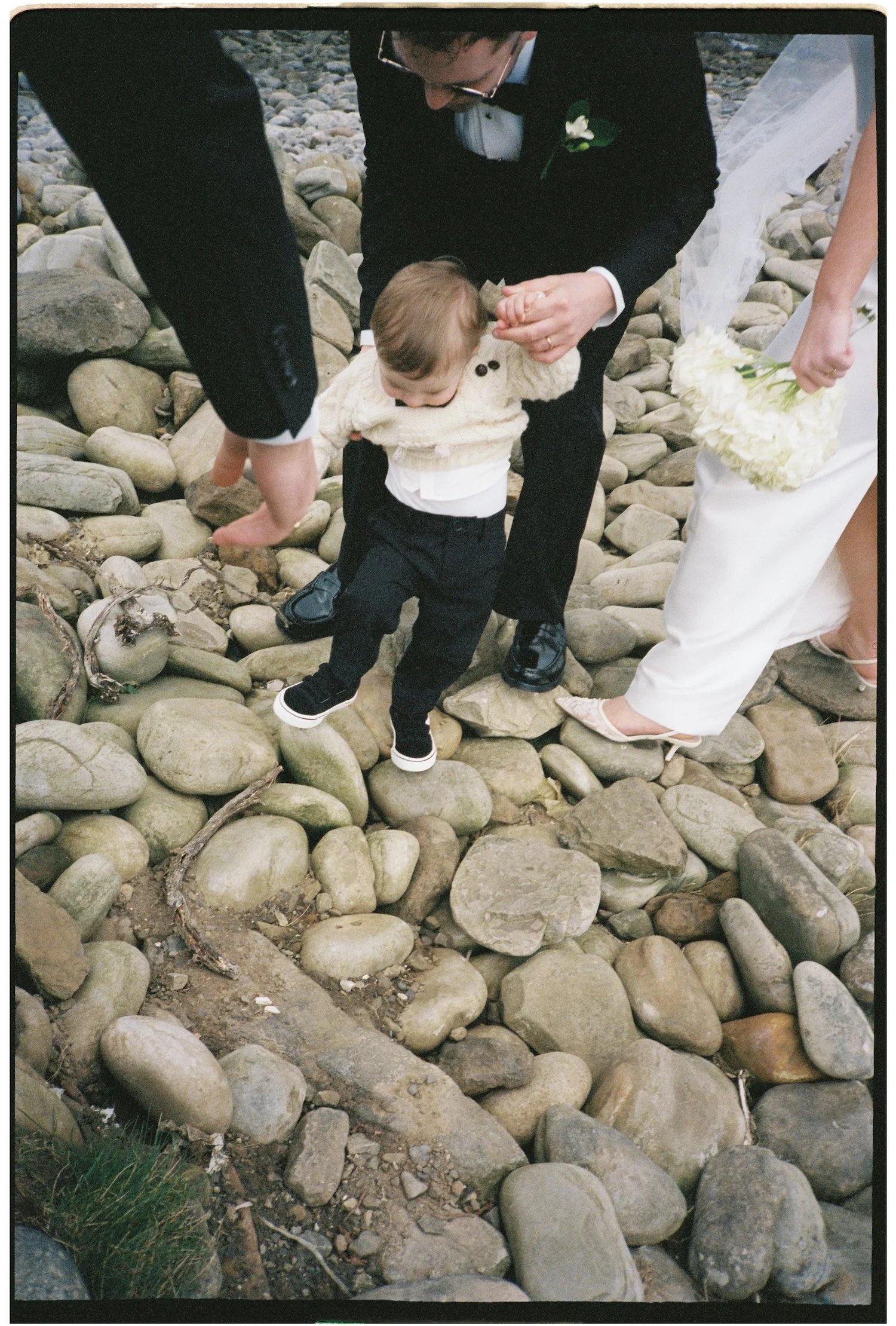 A man dressed in a tuxedo holding the hand of a young child on a rocky ground, with a woman holding a bouquet of white flowers nearby.