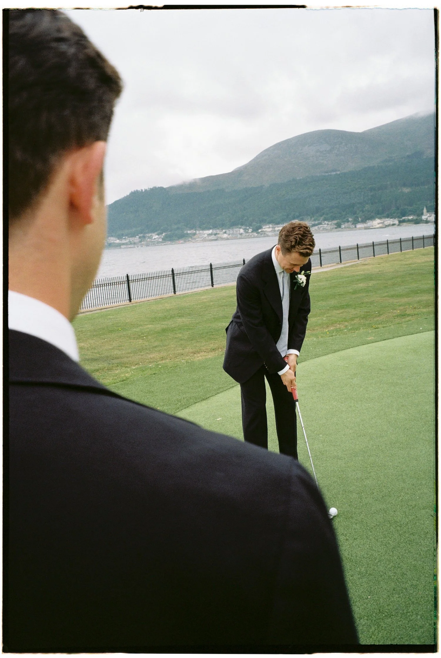 Two men dressed in suits on a golf course by a lake, with mountains in the background. One man is preparing to hit a golf ball.