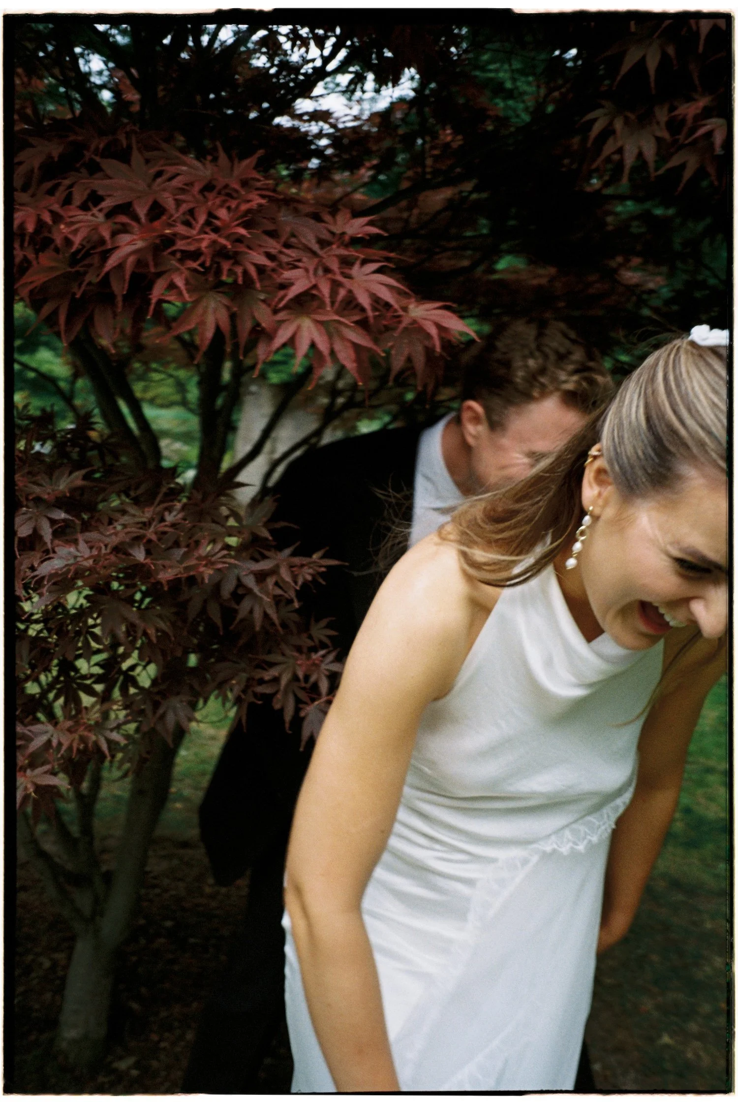 A woman in a white dress and pearl earrings standing in front of a person dressed in a tuxedo, partially visible, with a garden full of foliage and a red-leafed tree in the background.
