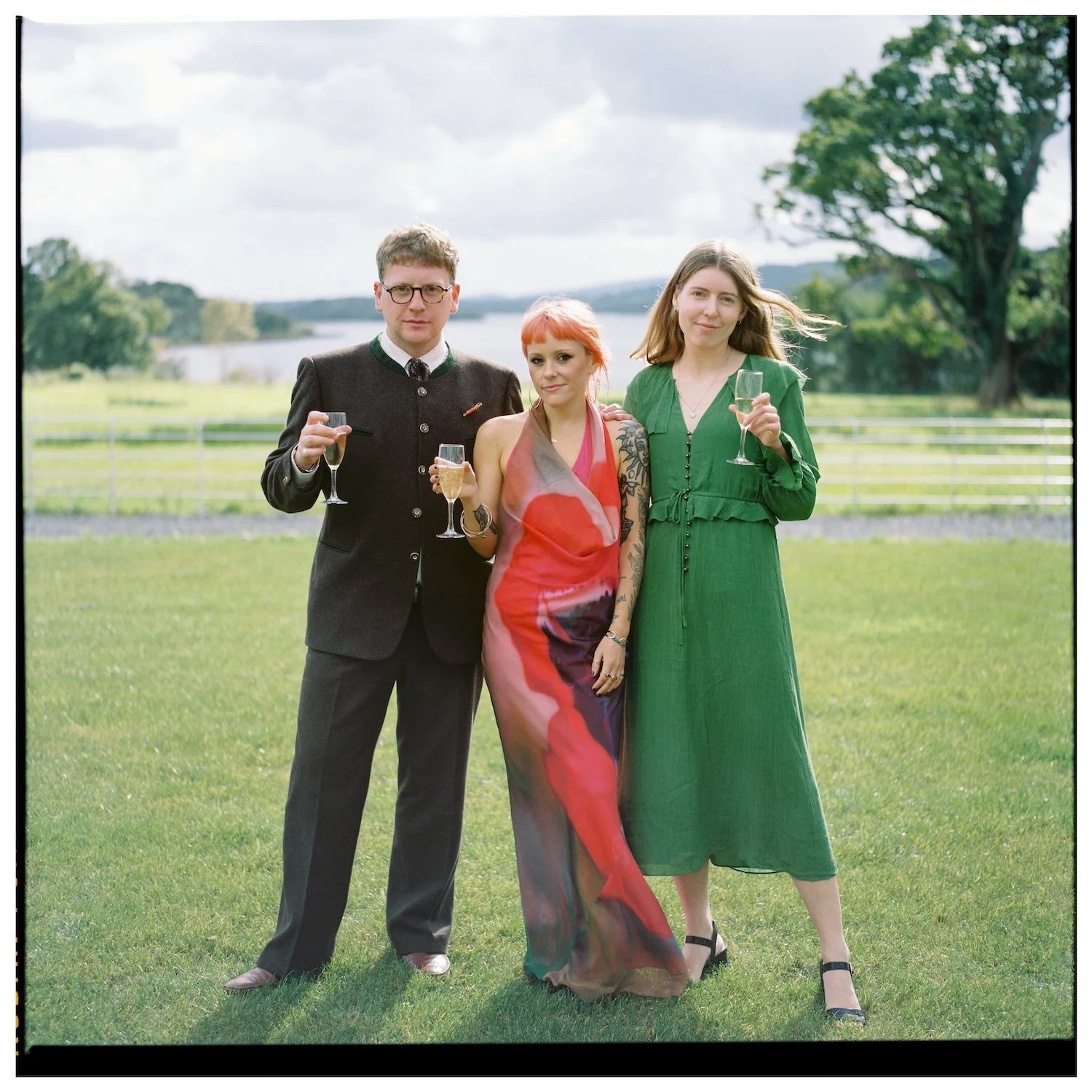 Three people standing outdoors in a grassy field, holding champagne glasses, with a large tree and lake in the background.