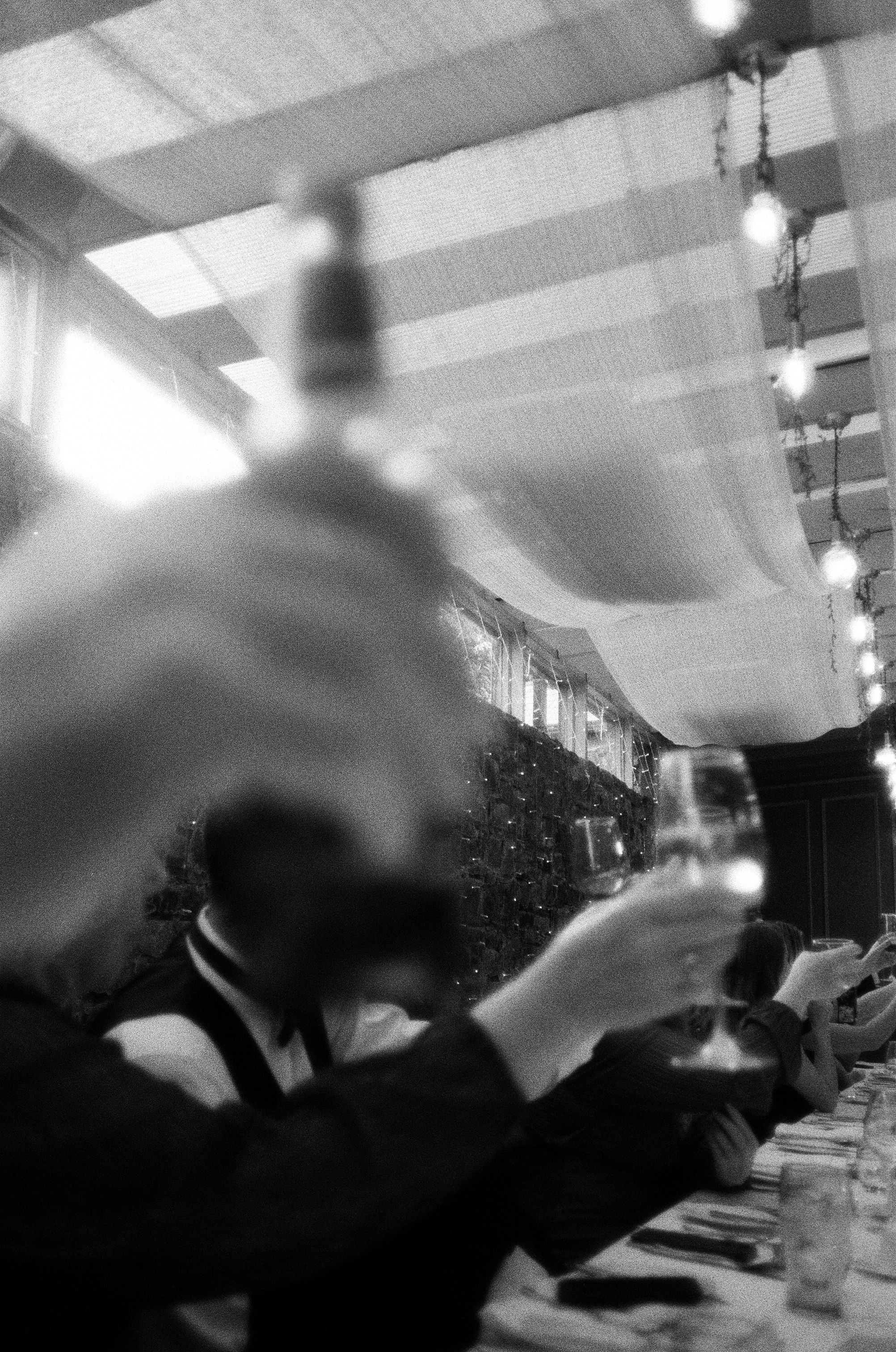 Black and white photo of people playing a piano, with one person playing and another watching, in a room decorated with string lights and a ceiling with visible grid pattern. The photo is blurry.