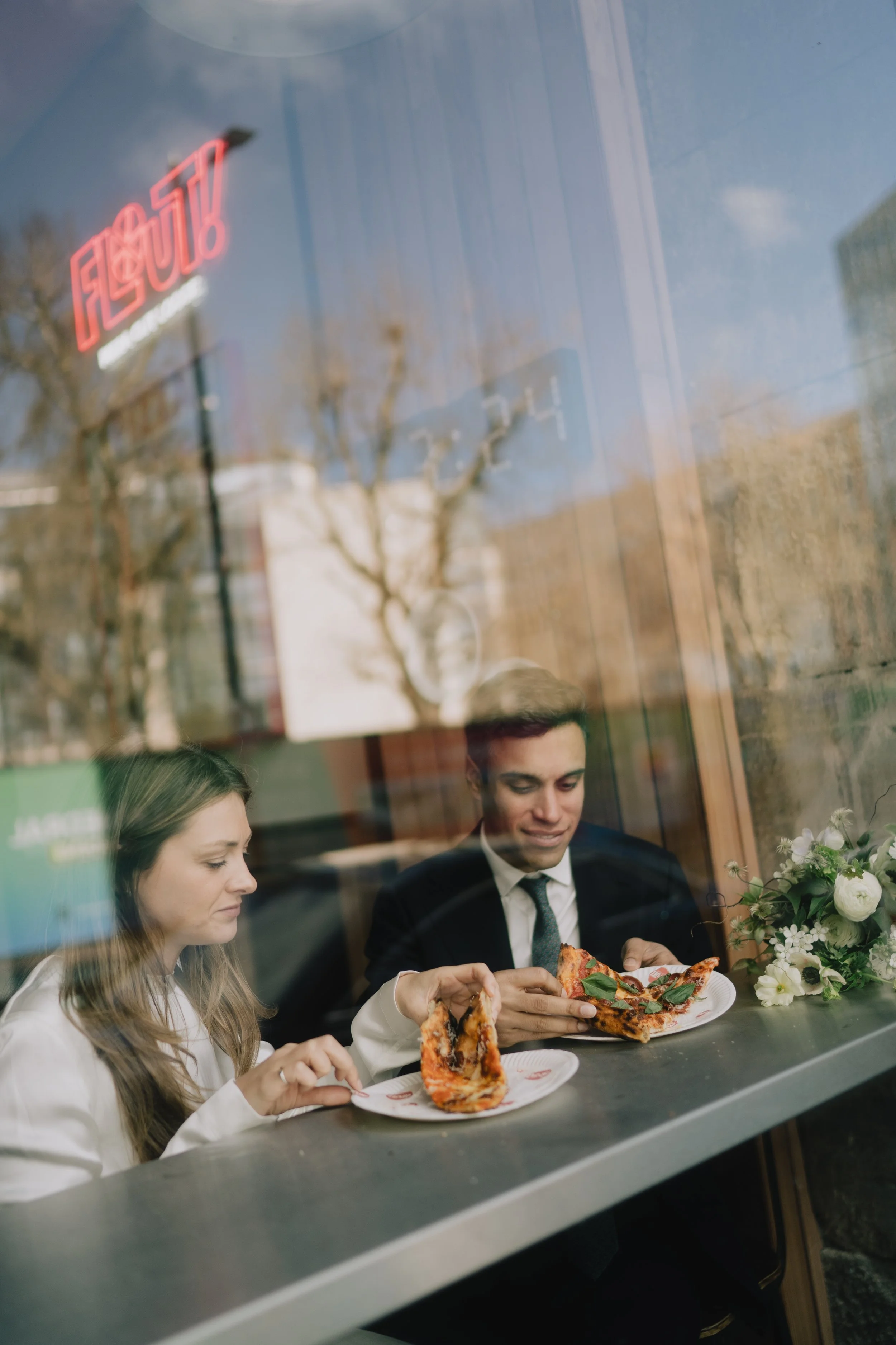 Two people, a woman with long hair and a man with short hair, are sitting at a table in a restaurant, eating pizza. The window reflects trees and a blue sky outside.