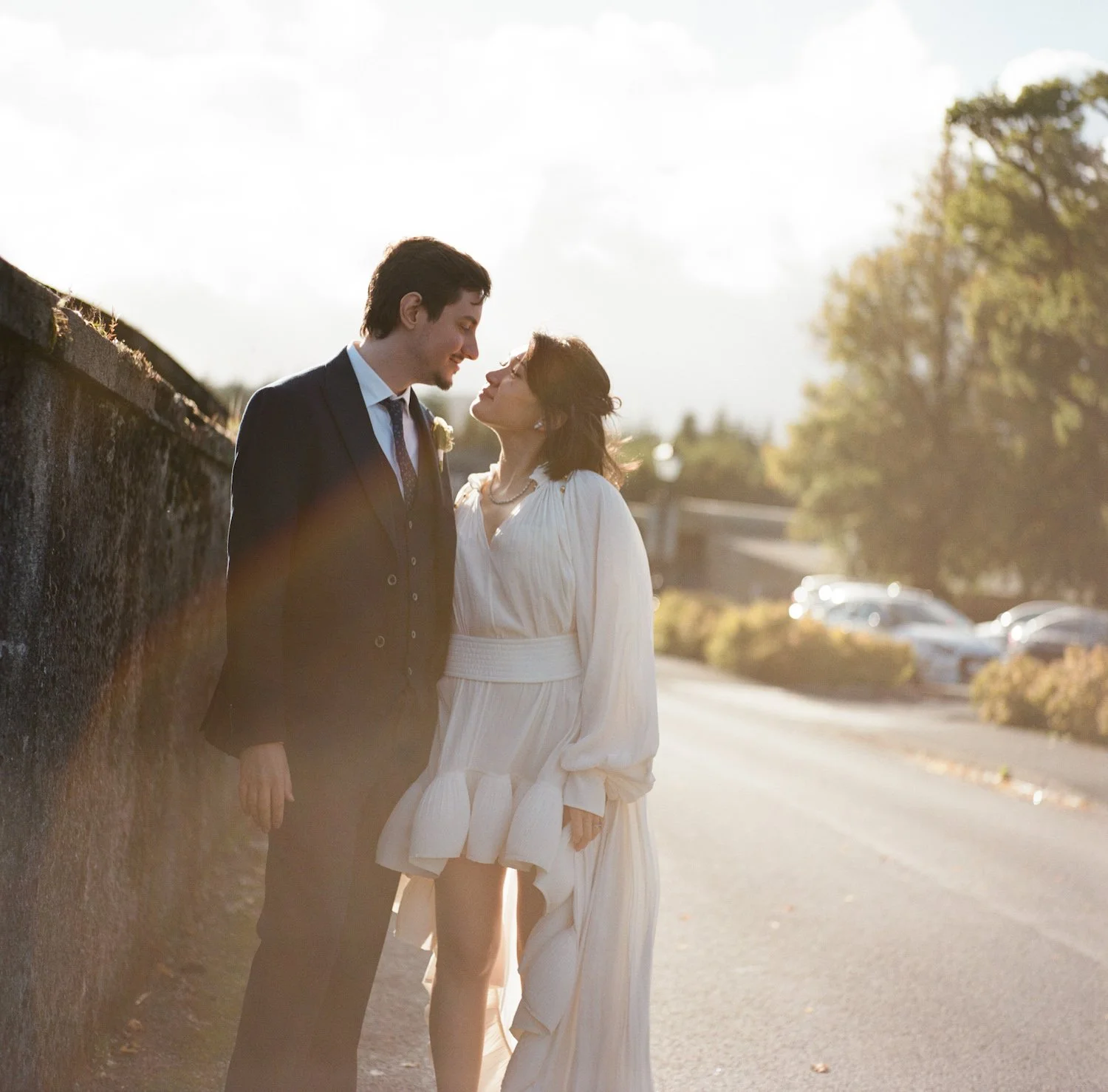 A bride and groom sharing a tender moment outdoors during sunset, with the groom in a suit and the bride in a white dress.