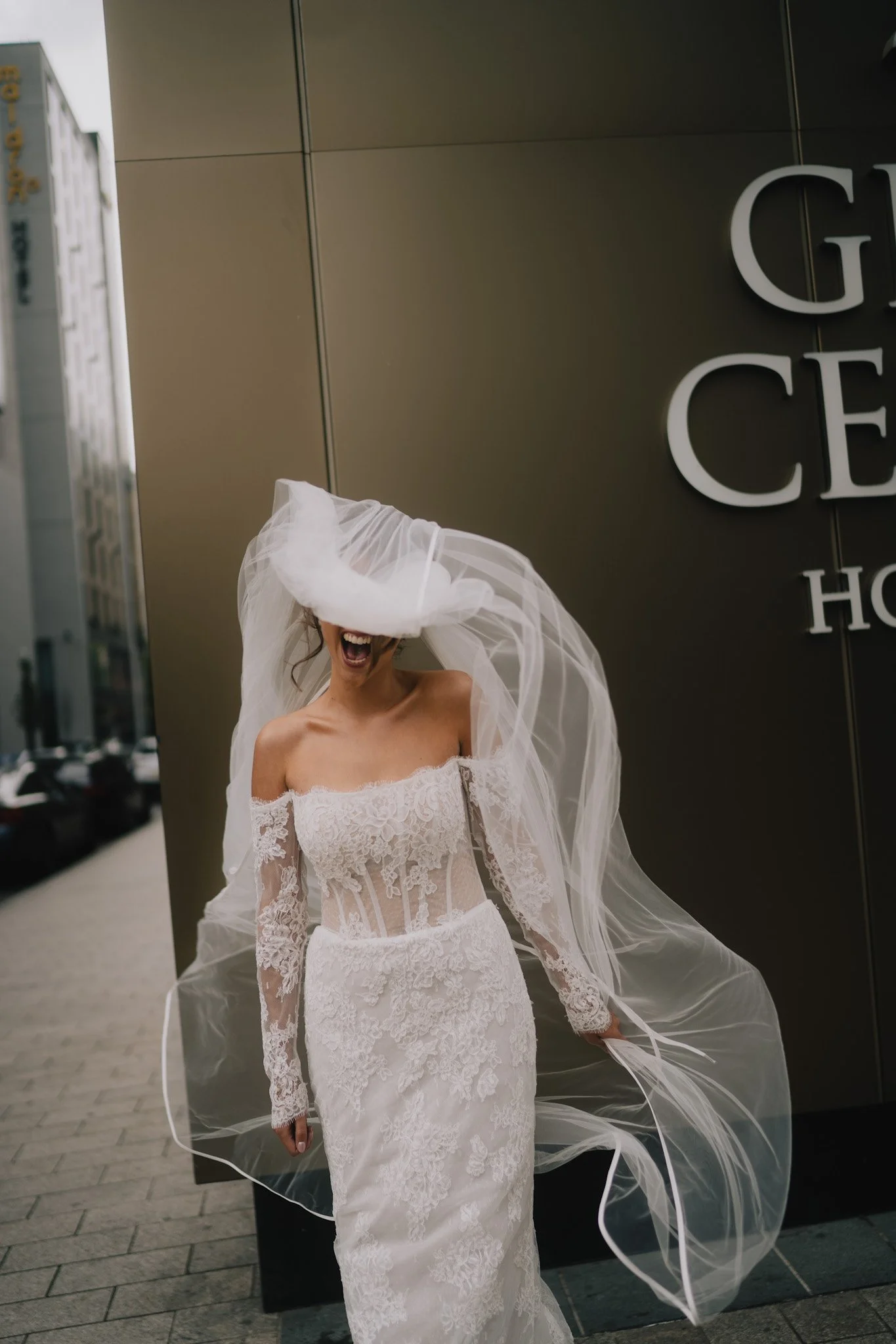 A woman in a white lace wedding dress with a sheer veil covering her face, standing outdoors near a building with dark panels and large letters, smiling with her mouth open.