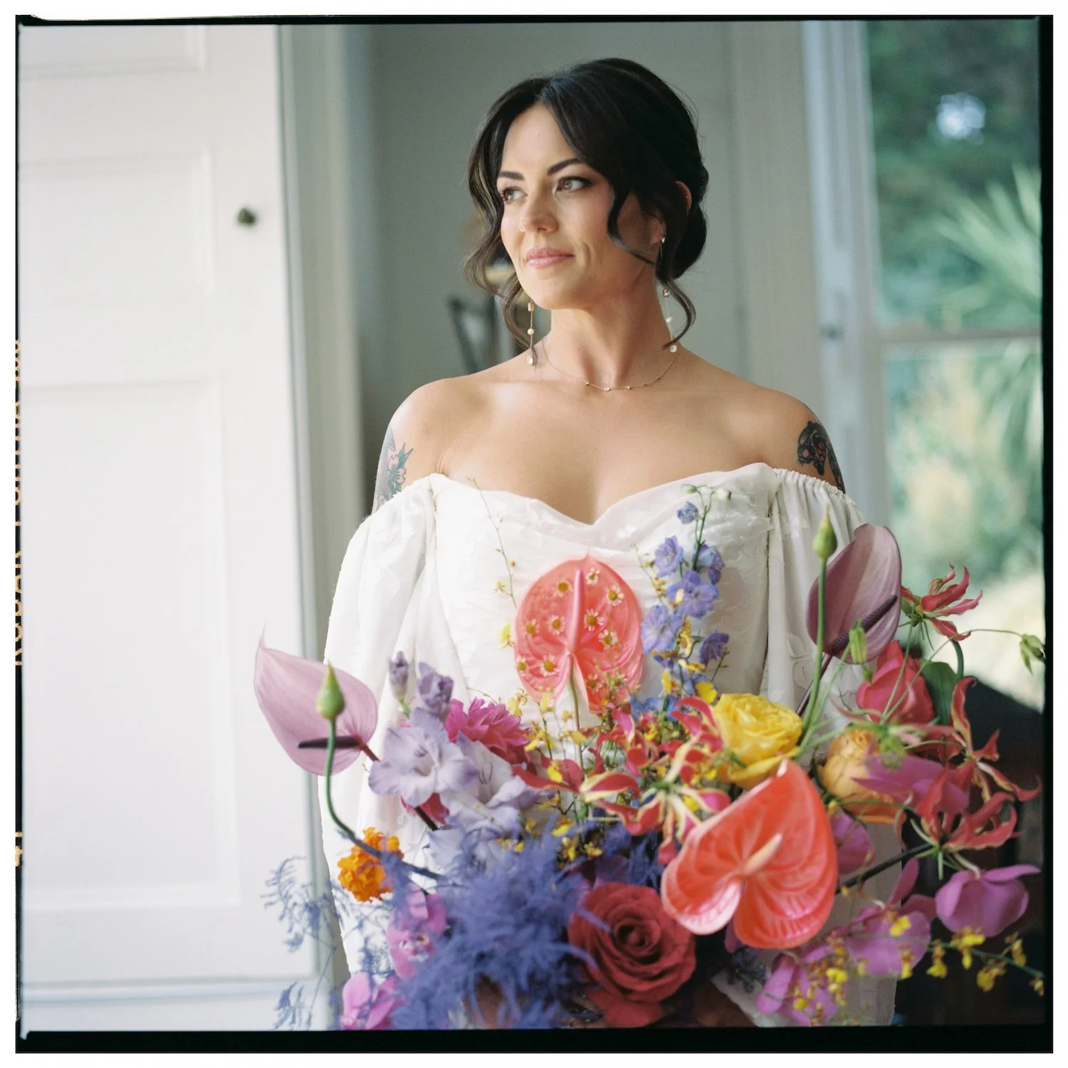 A woman with dark hair, tattoos, wearing a white off-the-shoulder dress, stands indoors holding a large colorful flower bouquet, with a window in the background.