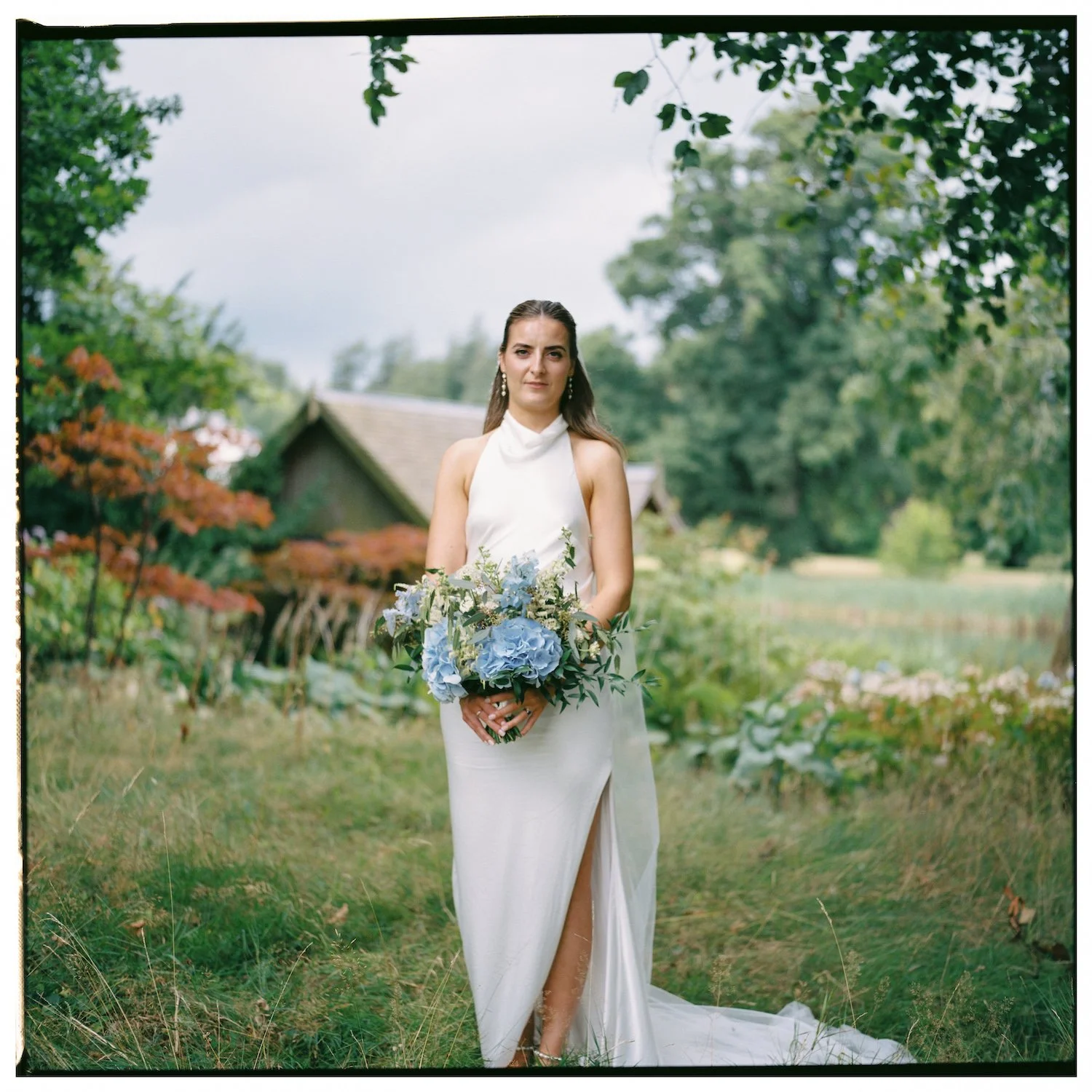 A woman in a white wedding dress holding a bouquet of blue hydrangeas standing in a garden with trees and a small building in the background.