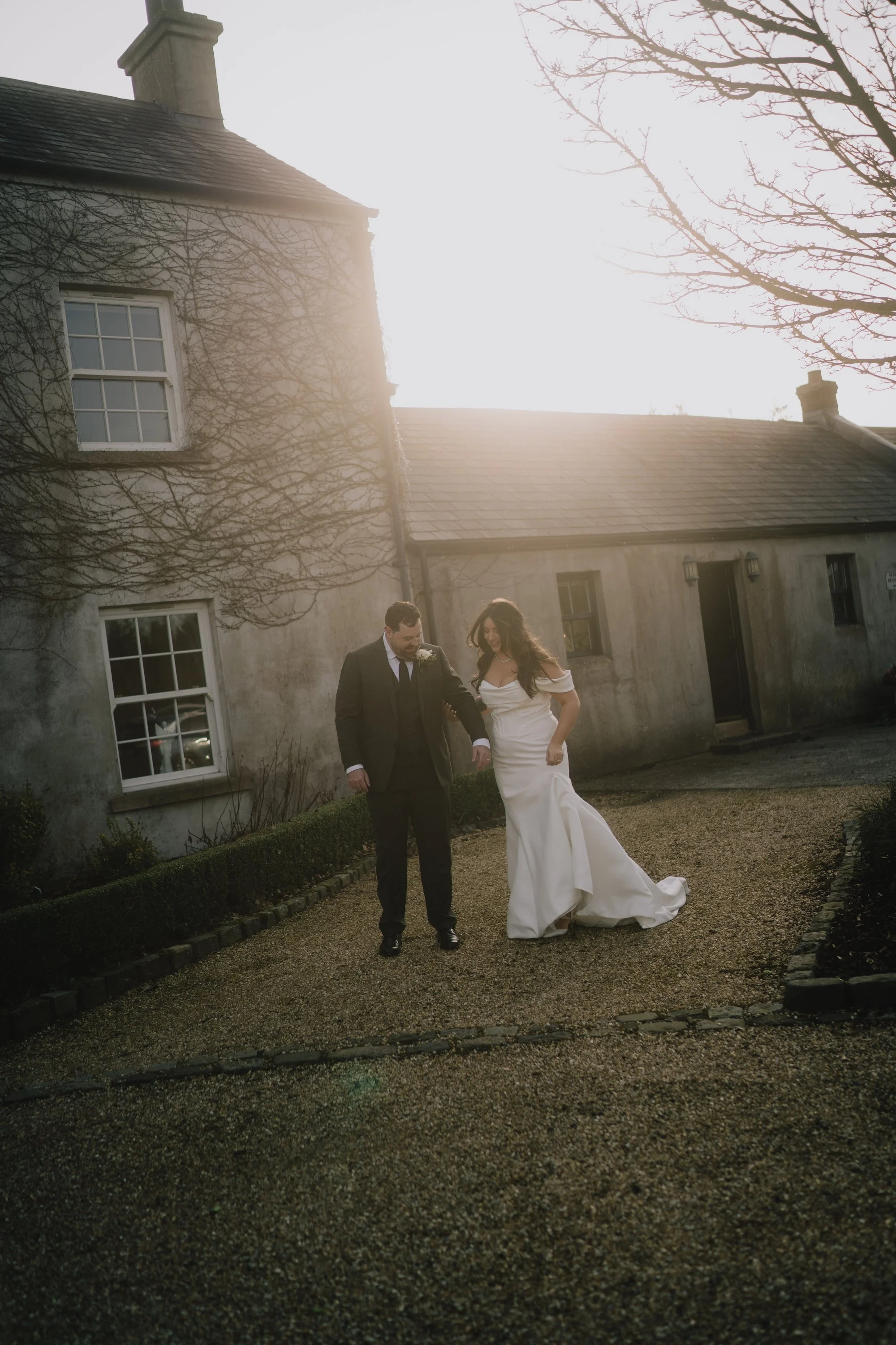 A bride and groom walking together outside near a stone building with leafless trees. The bride is wearing a white wedding dress and the groom is in a black suit. The sun is shining brightly behind the building, creating a backlight effect.