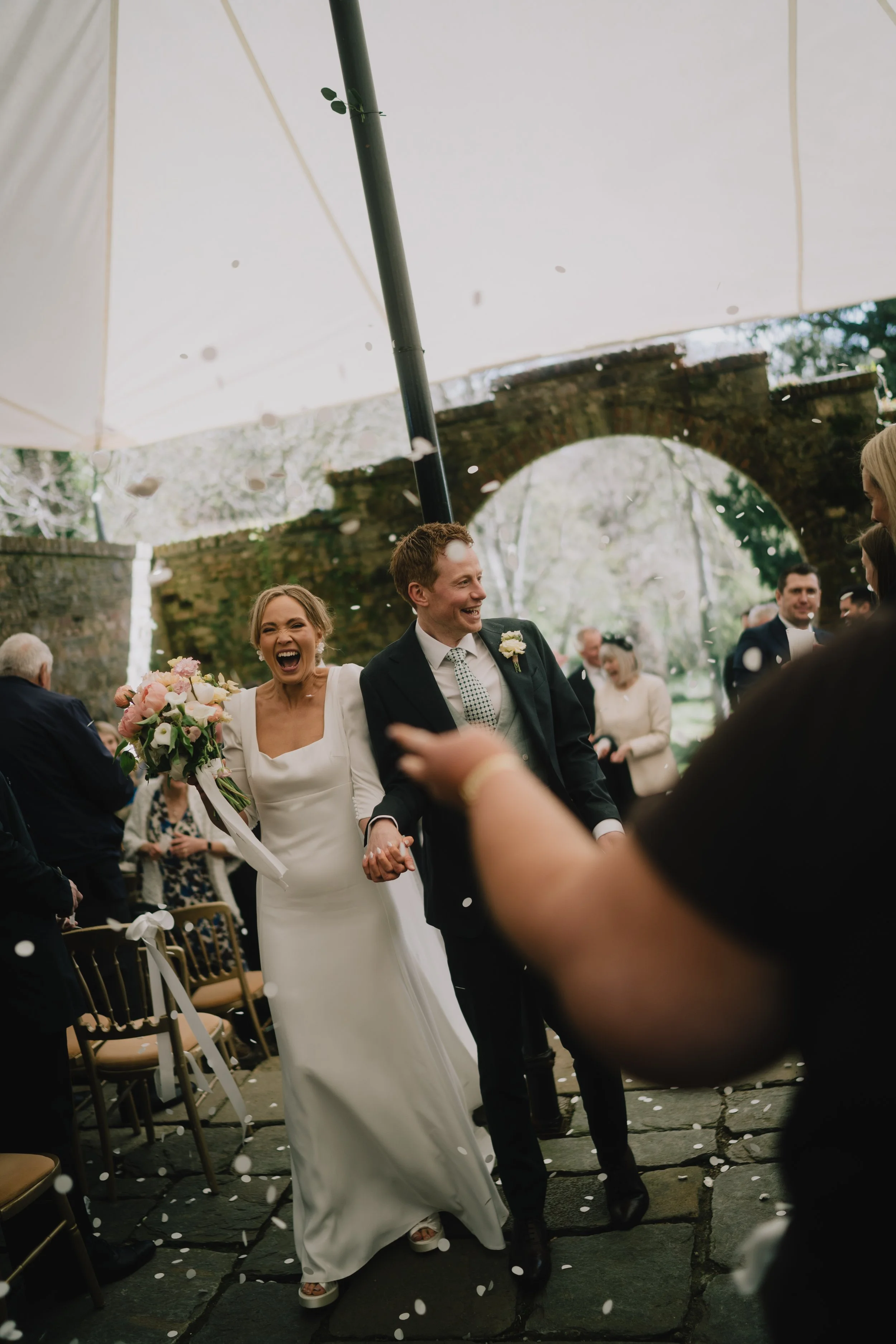 A bride and groom are smiling and holding hands while walking down an aisle at their wedding ceremony. The bride is wearing a white dress and holding a bouquet of pink and white flowers, and the groom is dressed in a black suit with a white shirt and
