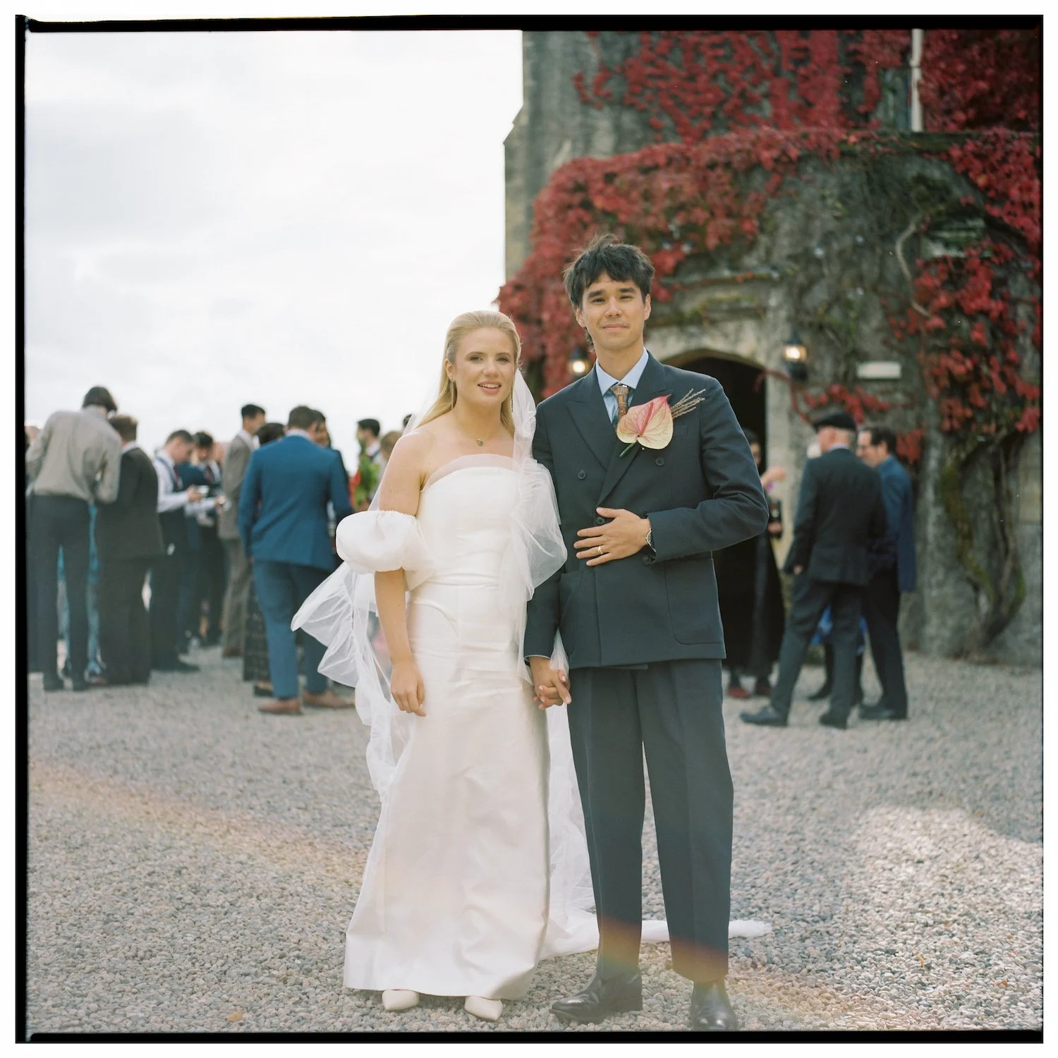 A newlywed couple holding hands and posing outdoors, with the bride in a white wedding dress and the groom in a dark suit with a boutonniere, in front of a stone building covered in red vine leaves, with other wedding guests in the background.