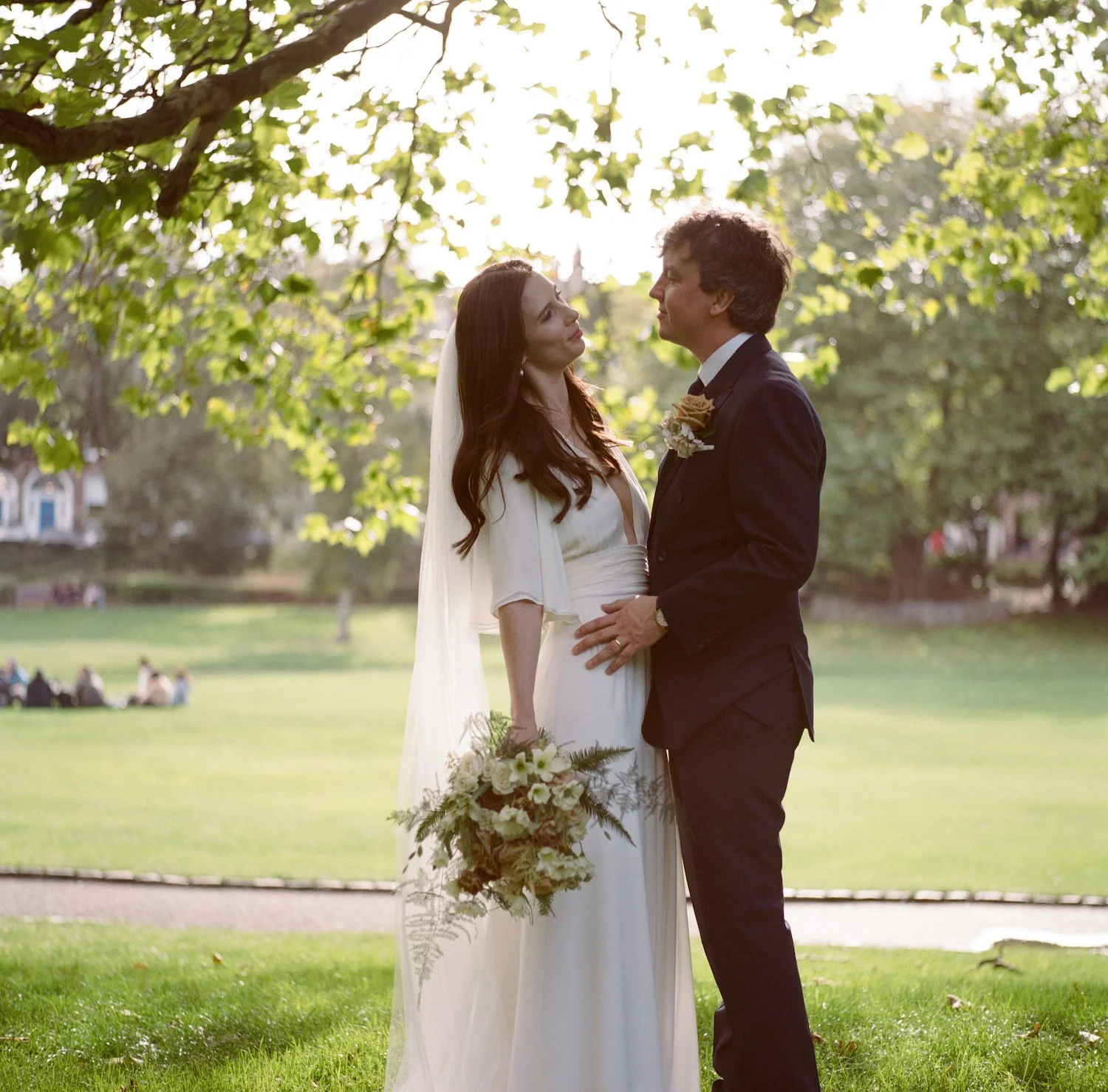 A bride and groom standing close together outdoors, with the bride holding a bouquet and wearing a white dress and veil, while the groom is in a dark suit with a boutonniere, looking into each other's eyes under a leafy tree.