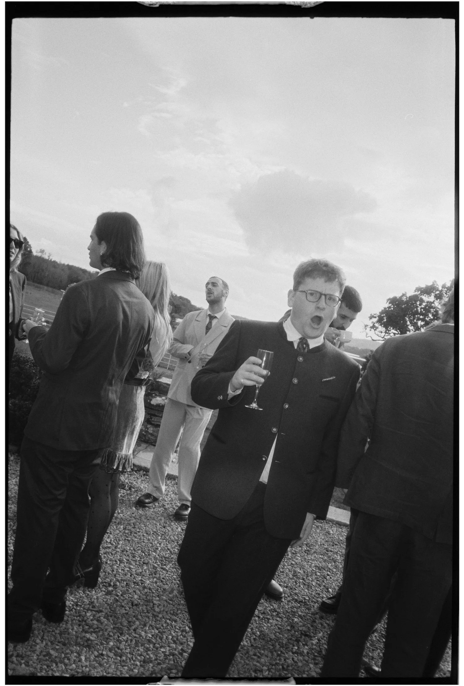 Group of people dressed in formal attire standing outdoors on a gravel surface, with some holding glasses, under a partly cloudy sky.