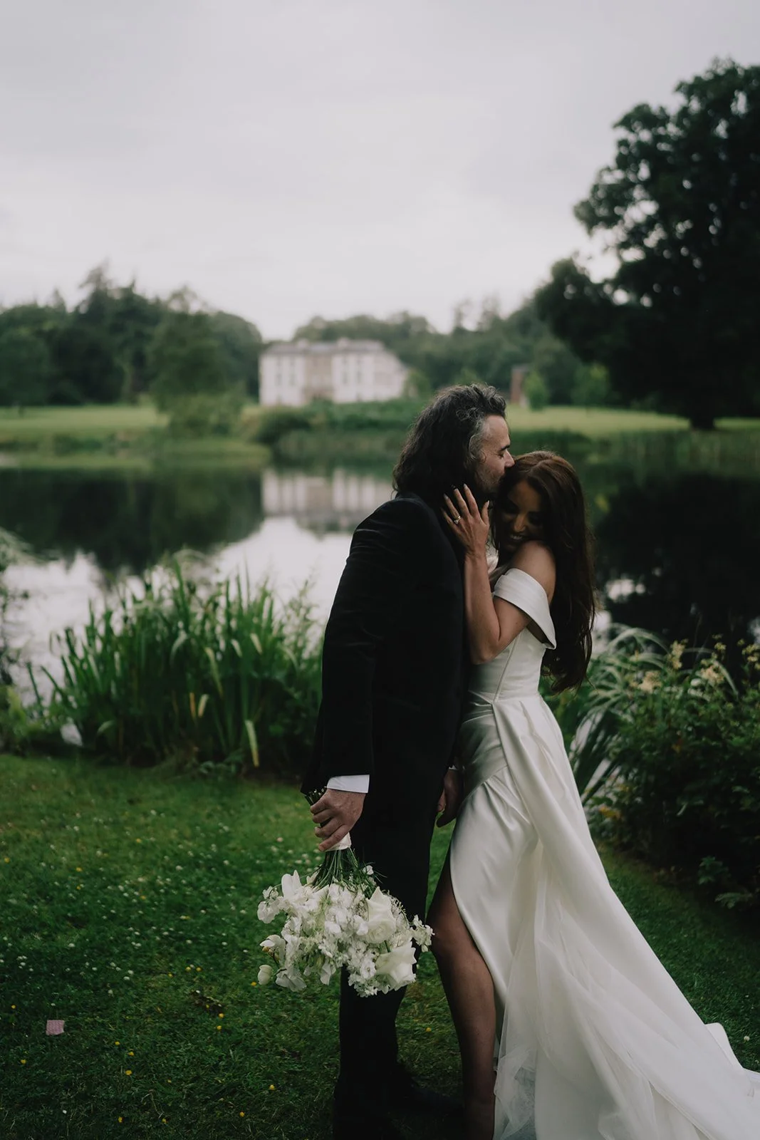 A couple on their wedding day, embracing and smiling by a pond in a lush green outdoor setting, with a large white house in the background.
