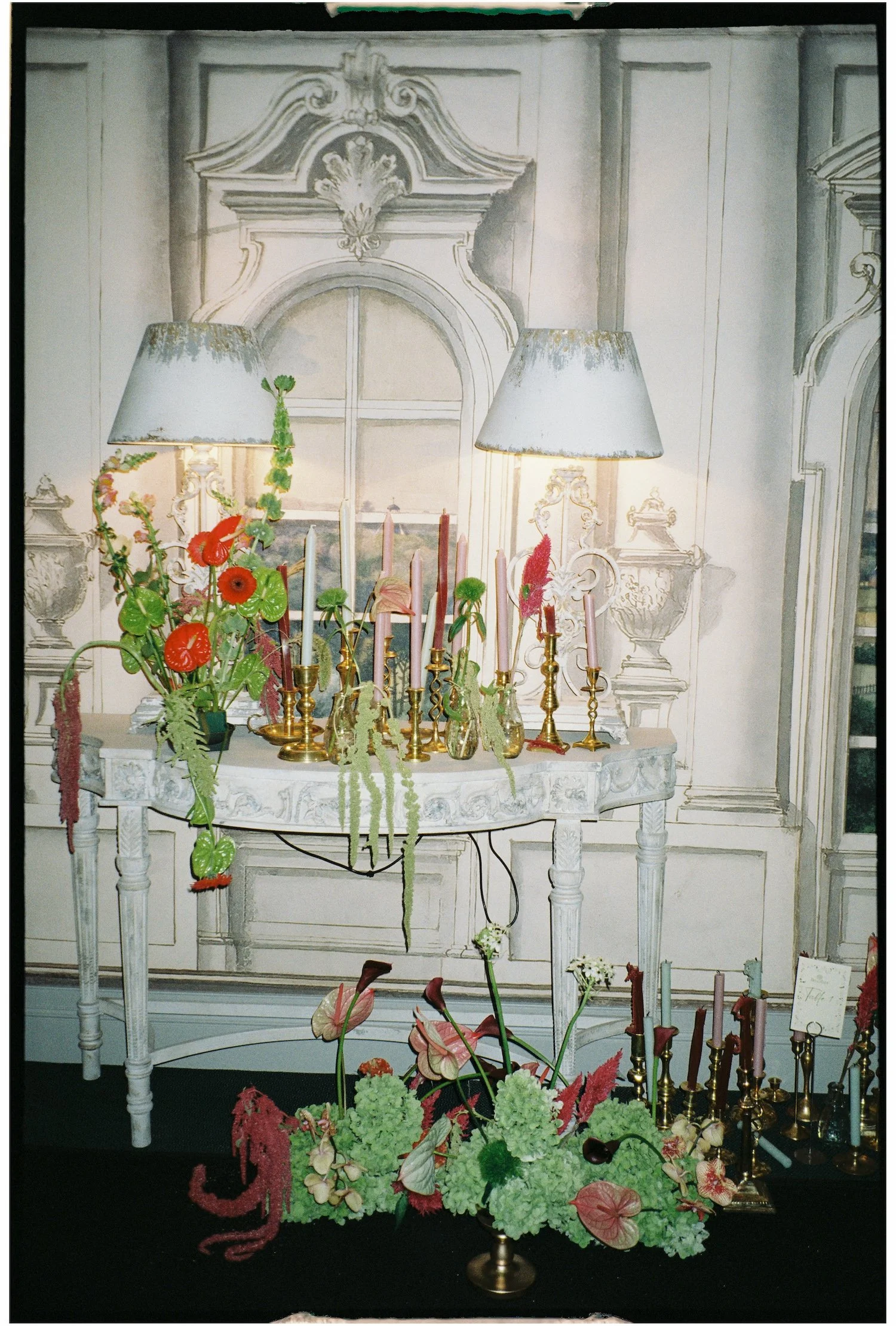 A floral arrangement with pink and green flowers and foliage on a white table in front of a background with ornate wall decorations, two lamps with distressed shades, and gold candlesticks with pink and red candles.